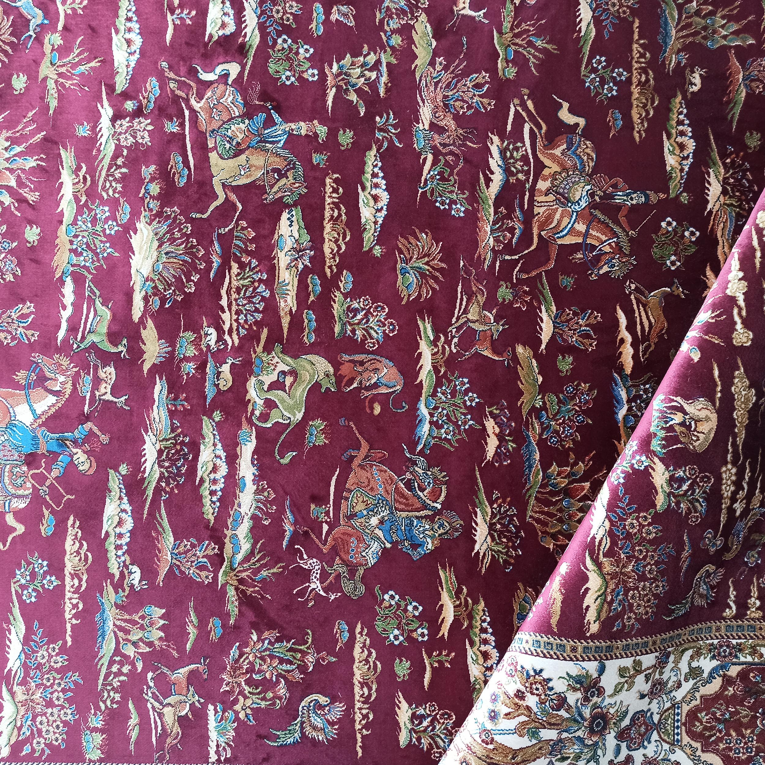 Tapis bordeaux à motifs traditionnels détaillés, figuratifs et floraux. Sa bordure repliée révèle un décor crème et bleu