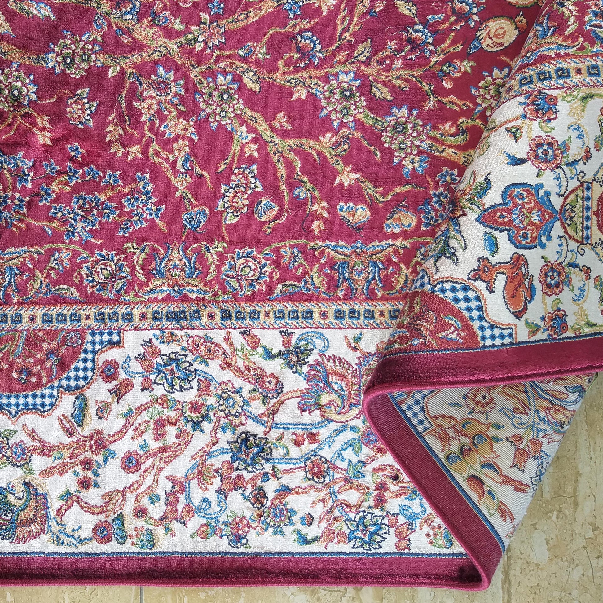 Tapis bordeaux orné de motifs floraux et de lianes multicolores. Sa bordure crème à motifs floraux et géométriques bleus et rouges est visible, le tapis étant partiellement roulé