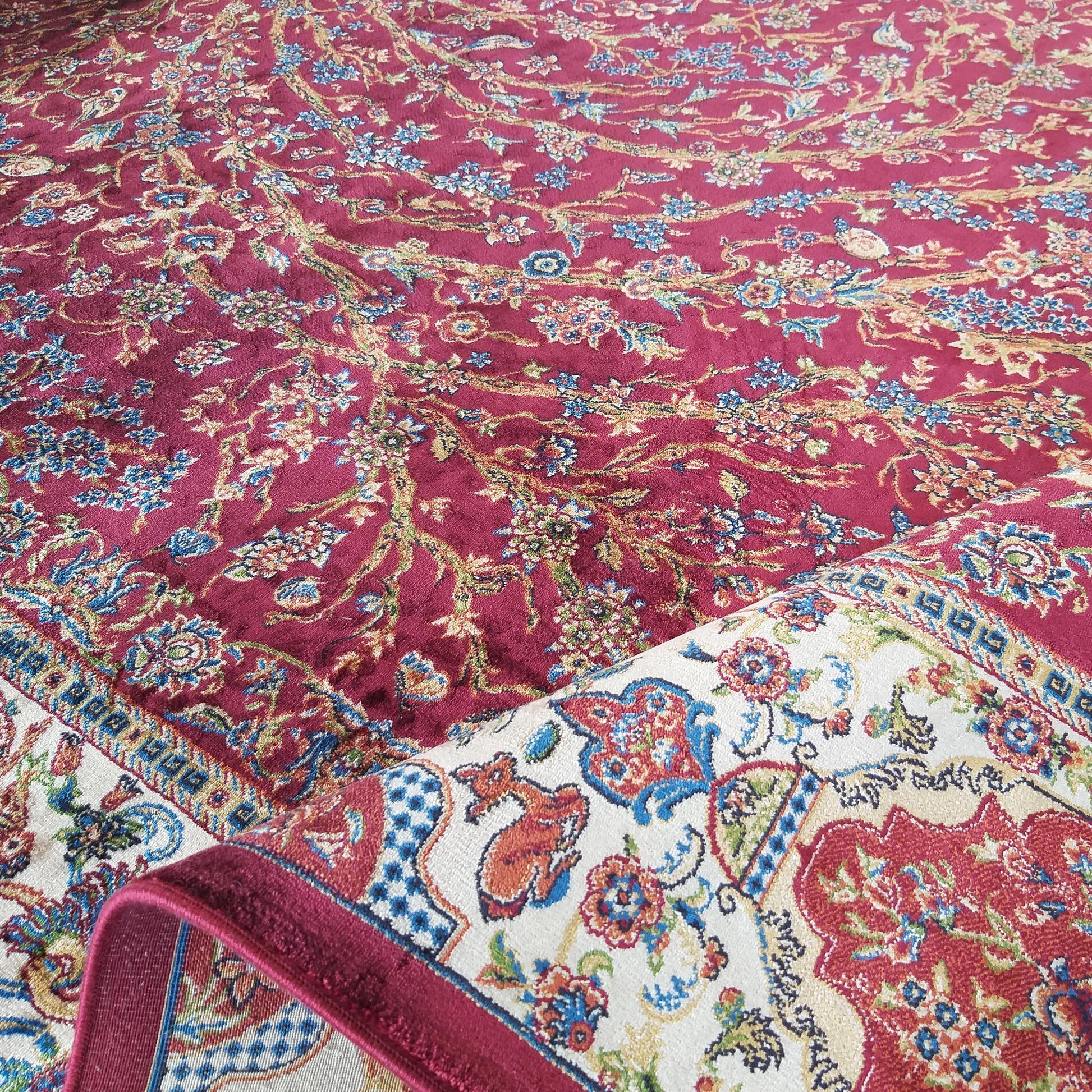 Tapis bordeaux aux motifs floraux et végétaux complexes en bleu, vert et beige, partiellement enroulé dévoilant une bordure crème