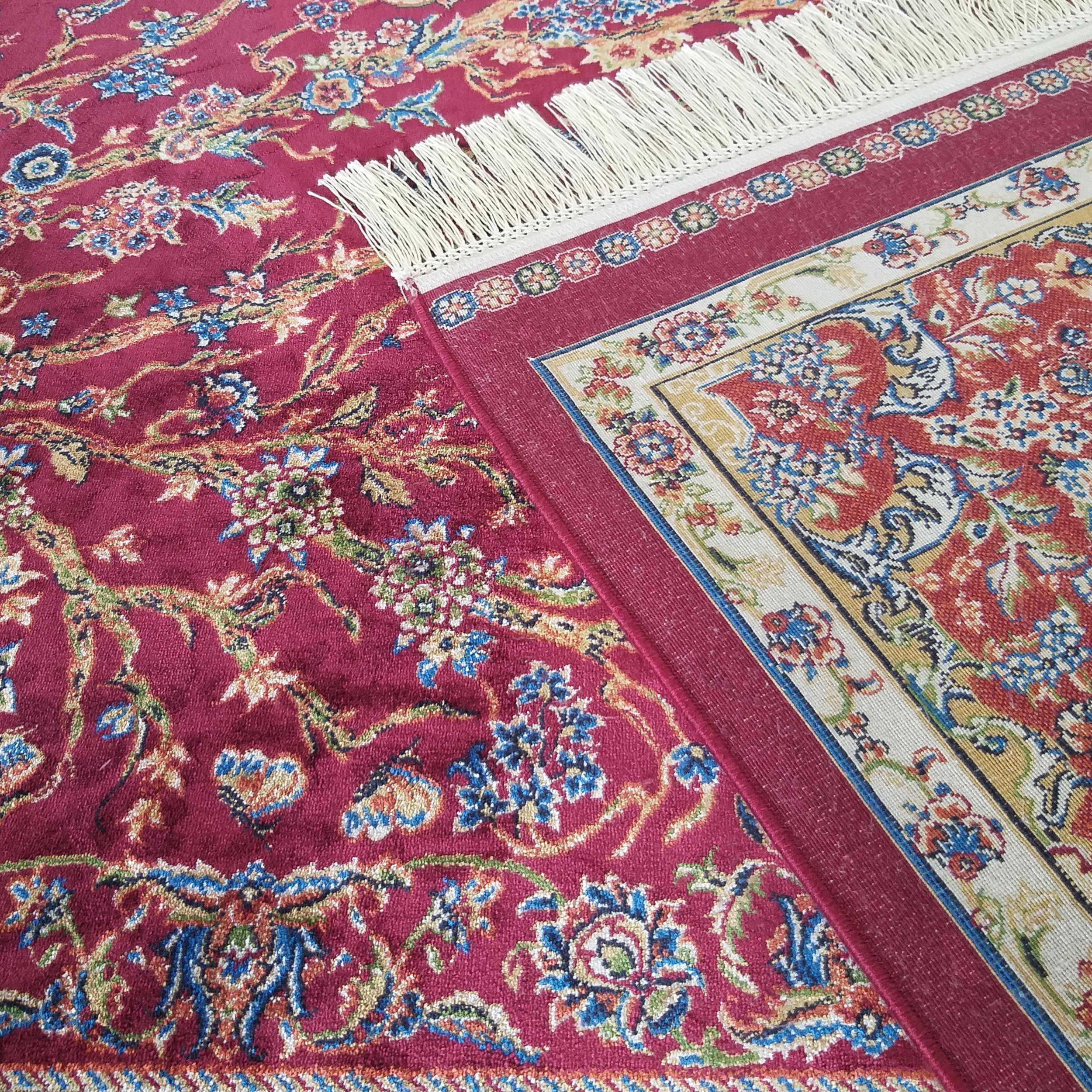 Tapis textile rouge bordeaux avec un motif floral ornemental en bleu, or et vert. Des franges crème et une bordure travaillée sont apparentes