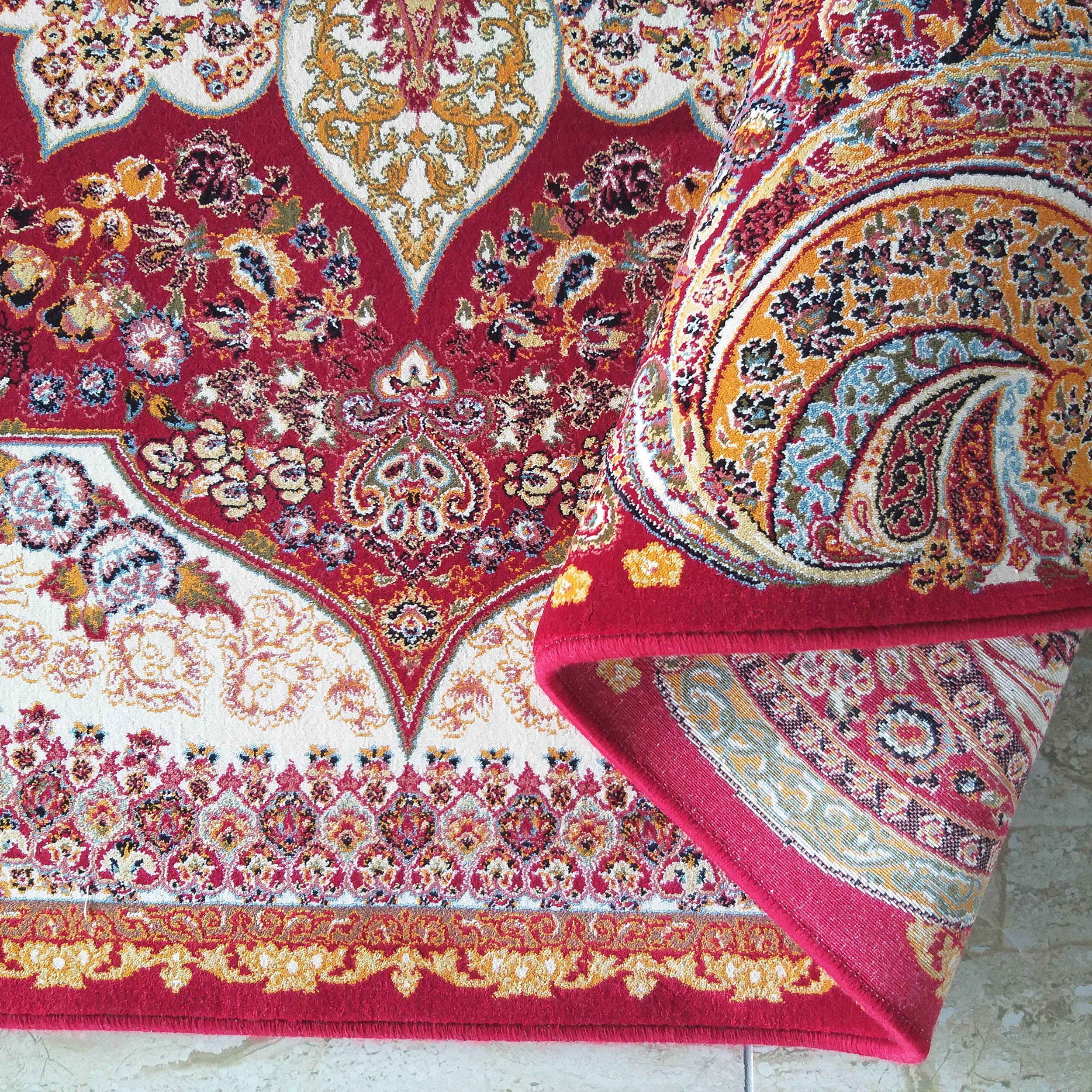 Grand tapis aux motifs orientaux complexes rouges, blancs et dorés. Une section est enroulée, révélant la richesse de son dessin
