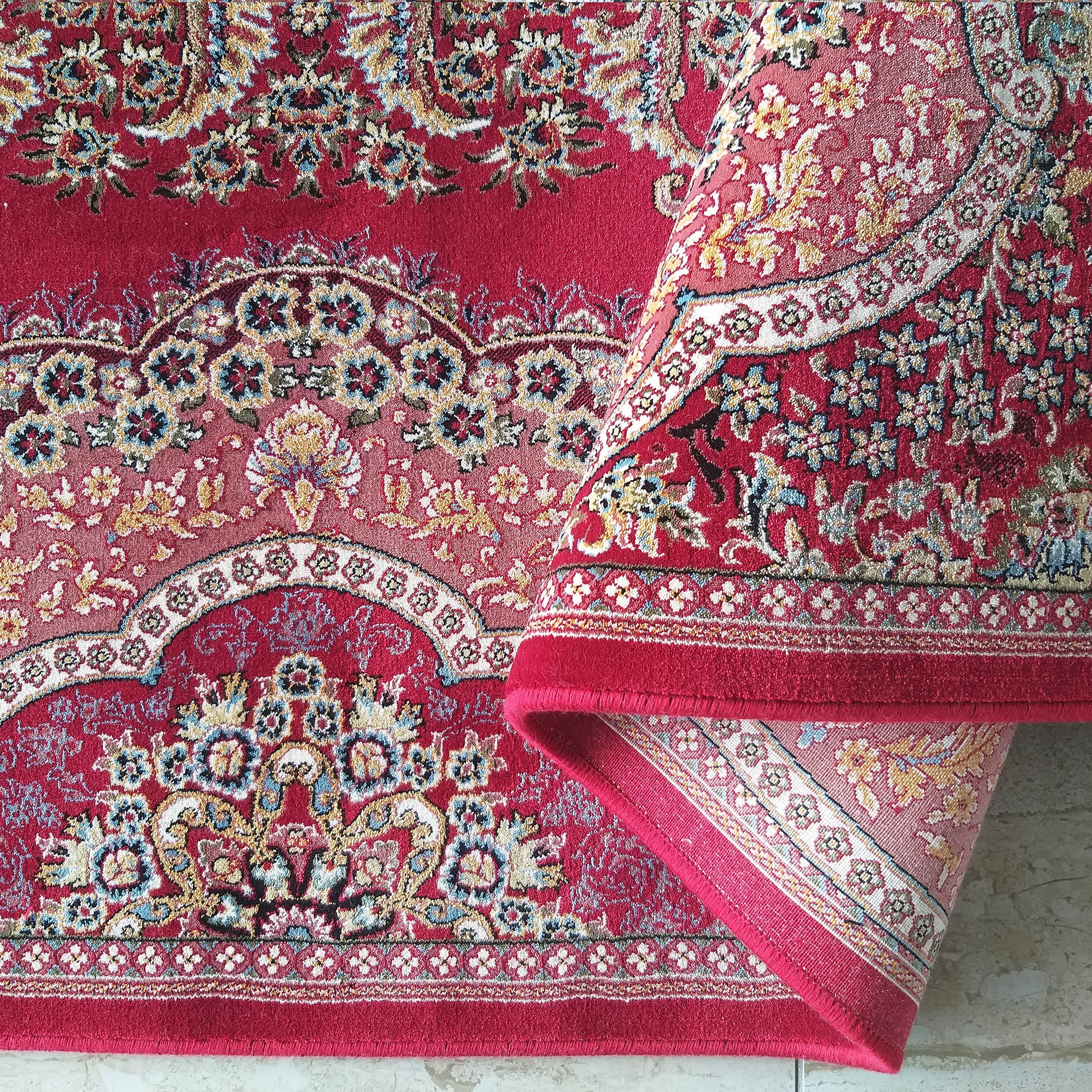 Tapis rouge grenat orné de riches motifs floraux et arabesques en bleu, or, crème et bordeaux, partiellement enroulé