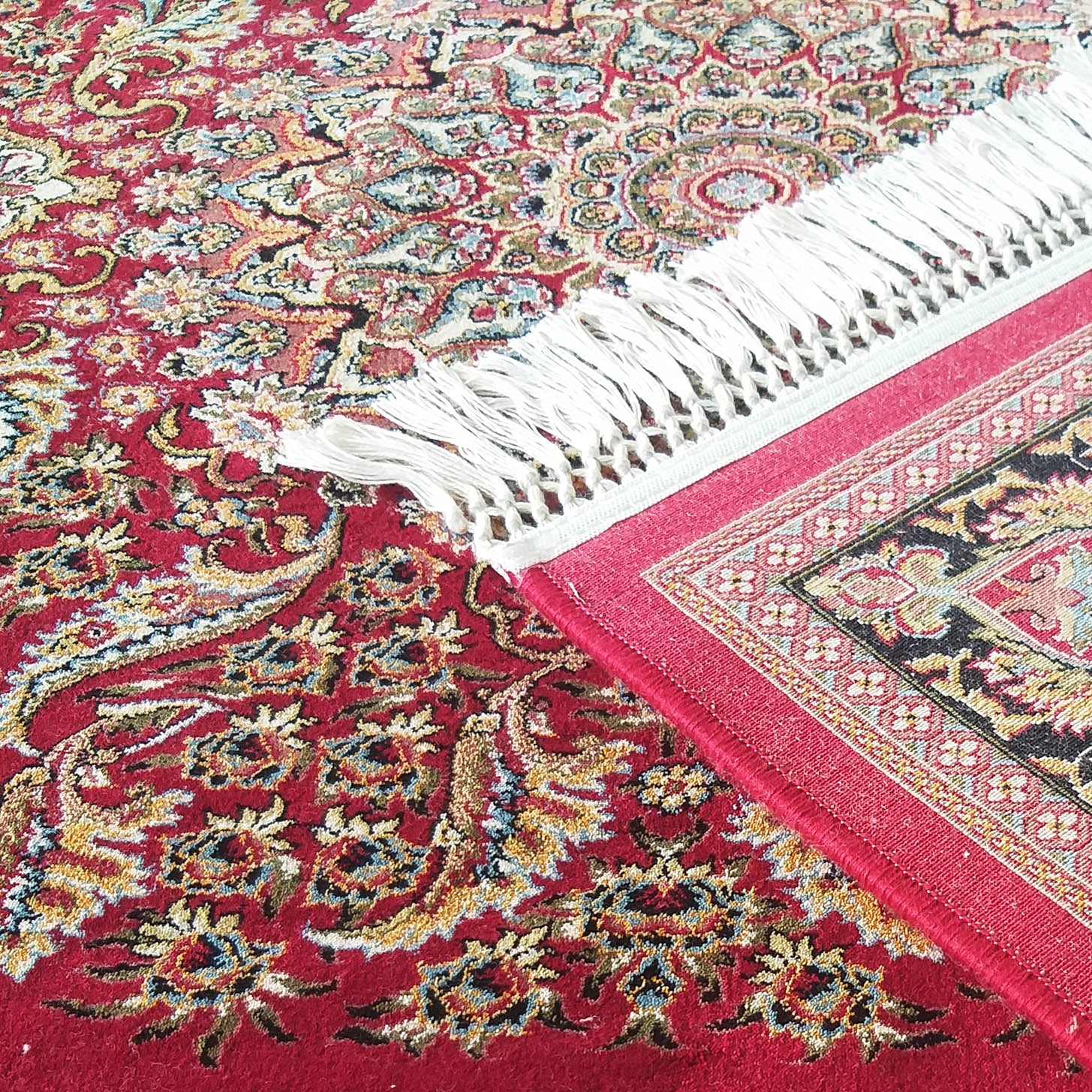 Tapis rouge orné de motifs floraux et végétaux complexes en doré, bleu, vert et crème. Des franges blanches décorent un bord du tapis