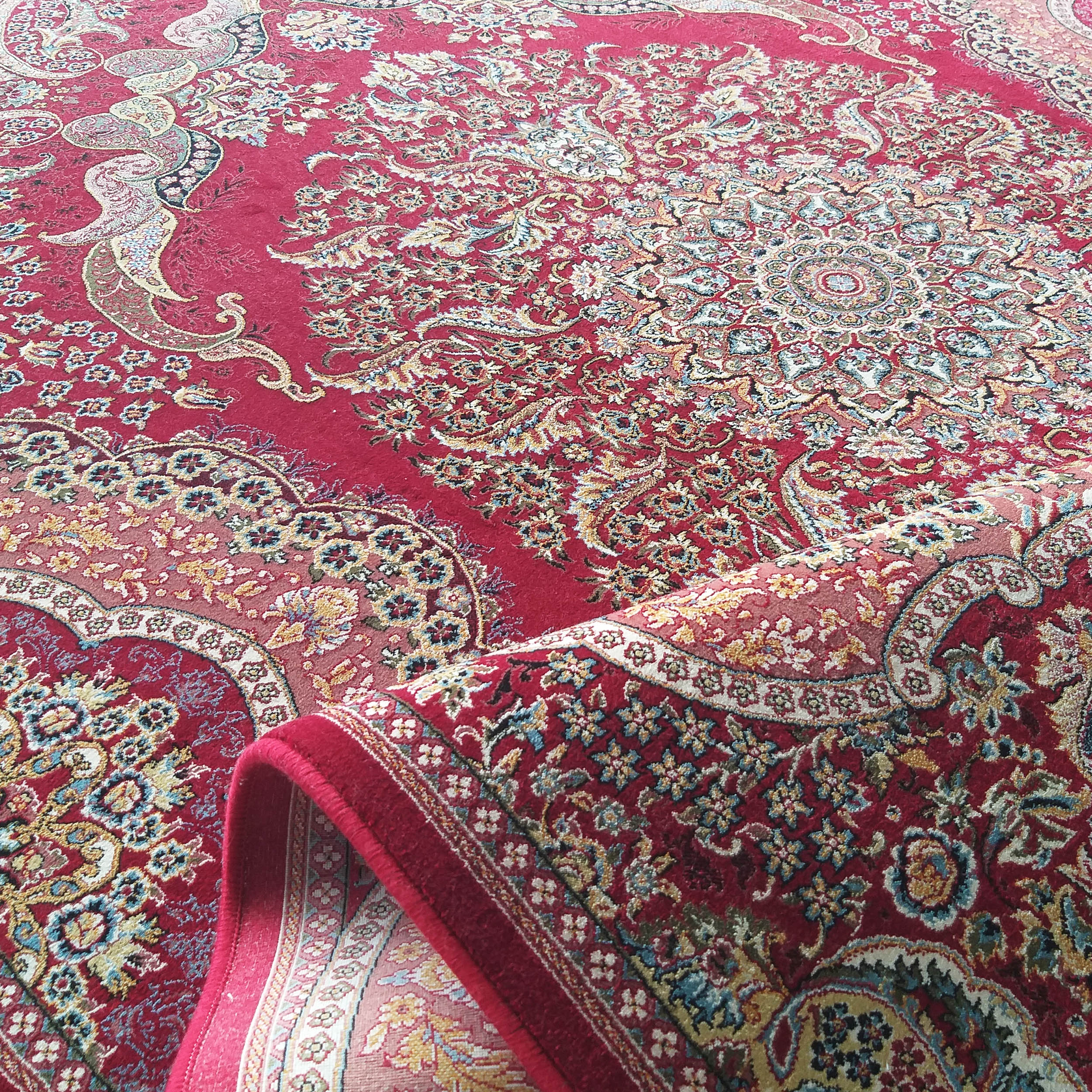 Tapis rouge orné de motifs floraux et cachemire complexes aux couleurs crème, or et bleues, partiellement roulé