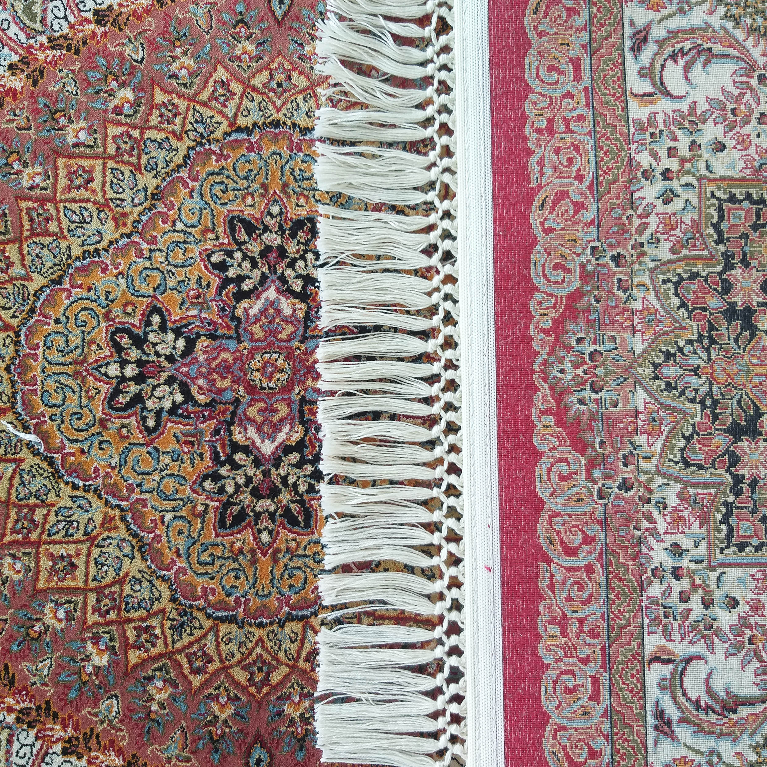 Tapis aux motifs floraux et arabesques complexes, dont les sections rouge et crème sont séparées par des franges blanches