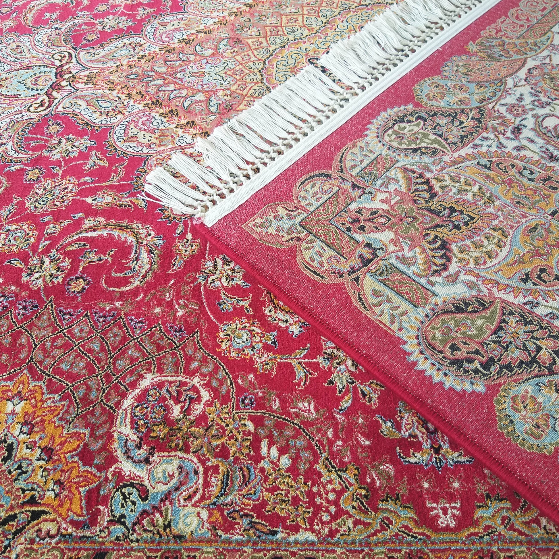 Deux tapis rouges superposés aux motifs orientaux complexes incluant fleurs et cachemires en bleu, or, crème et noir, le tapis supérieur présentant des franges blanches