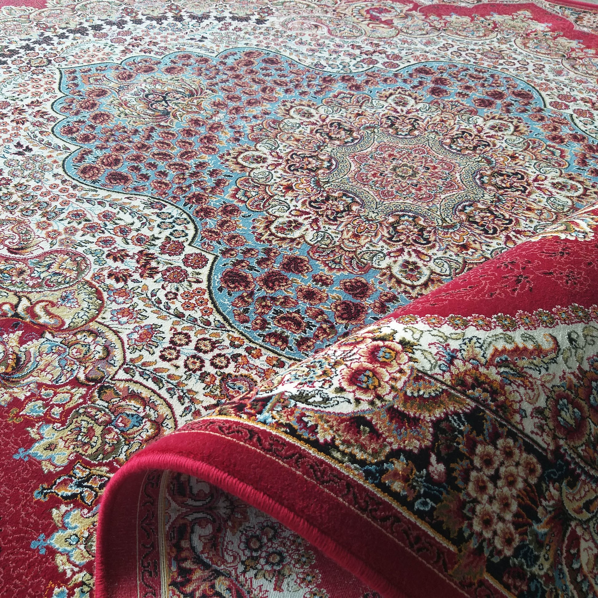 Tapis floral et à médaillon dans les tons rouge, crème et bleu clair, avec une bordure rouge vif. Une partie du tapis est enroulée