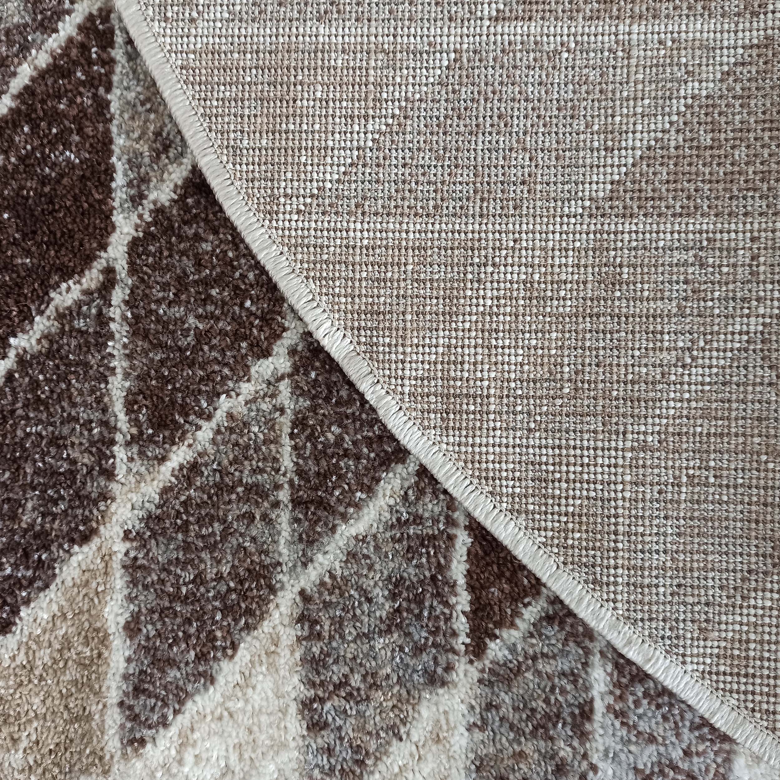 Tapis à motifs abstraits marron, beige et blanc cassé. Sa surface poilue et son envers tissé avec un bord surjeté sont visibles