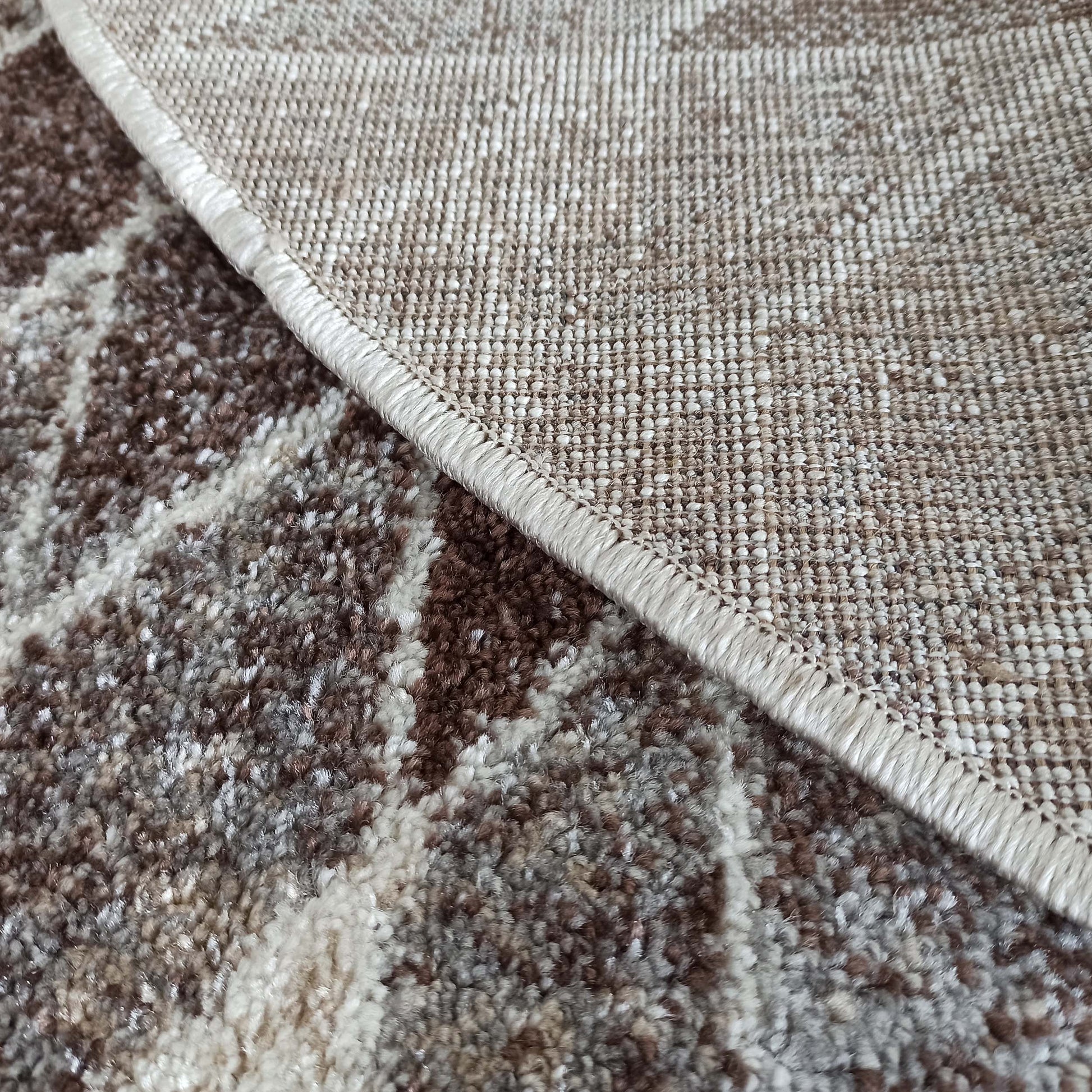 Tapis aux motifs abstraits marron, gris et blanc cassé, présentant sa texture moelleuse et son revers beige uni avec bord surjeté