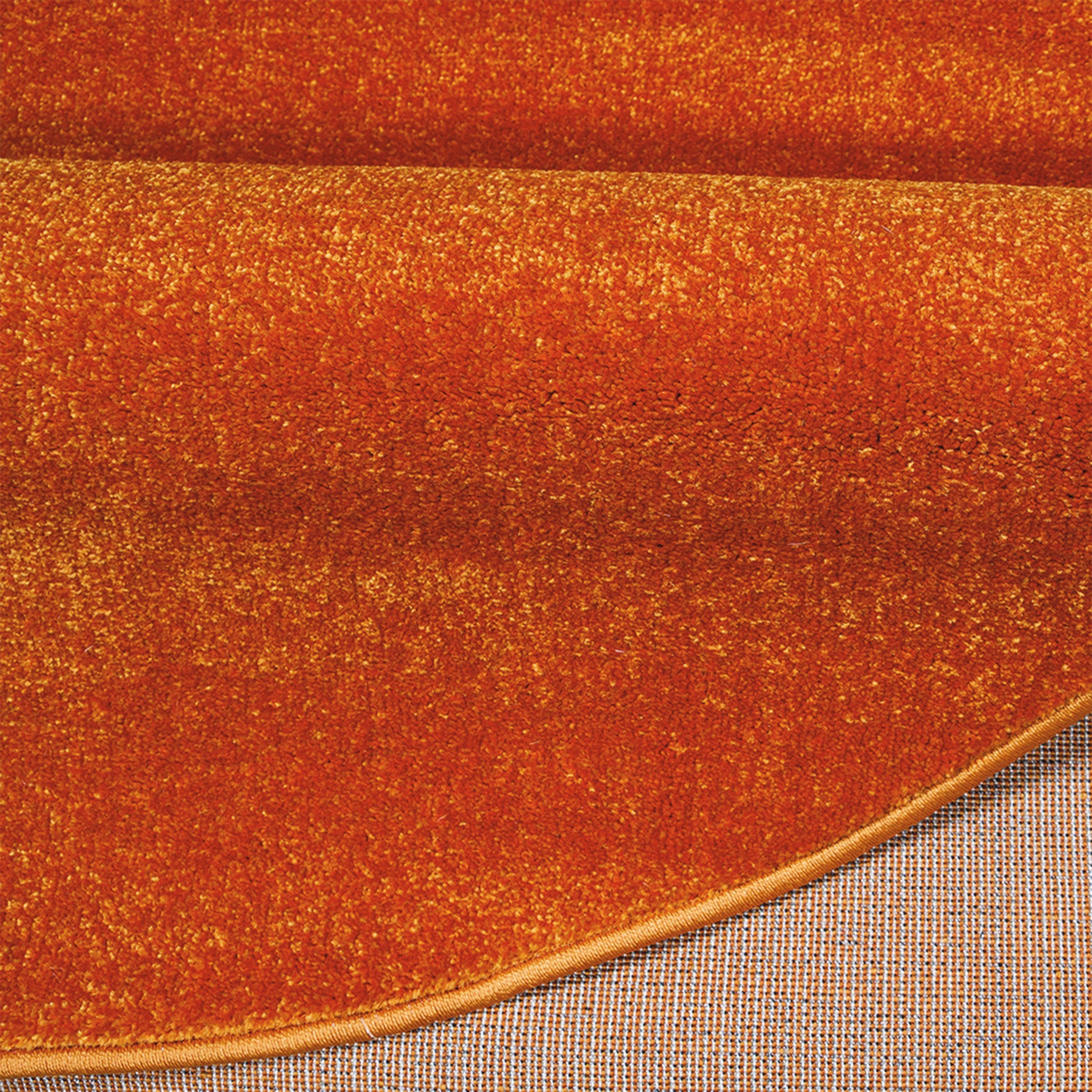 Tapis orange roux à la surface dense et moelleuse, partiellement replié, montrant son bord ourlé et son envers tissé quadrillé