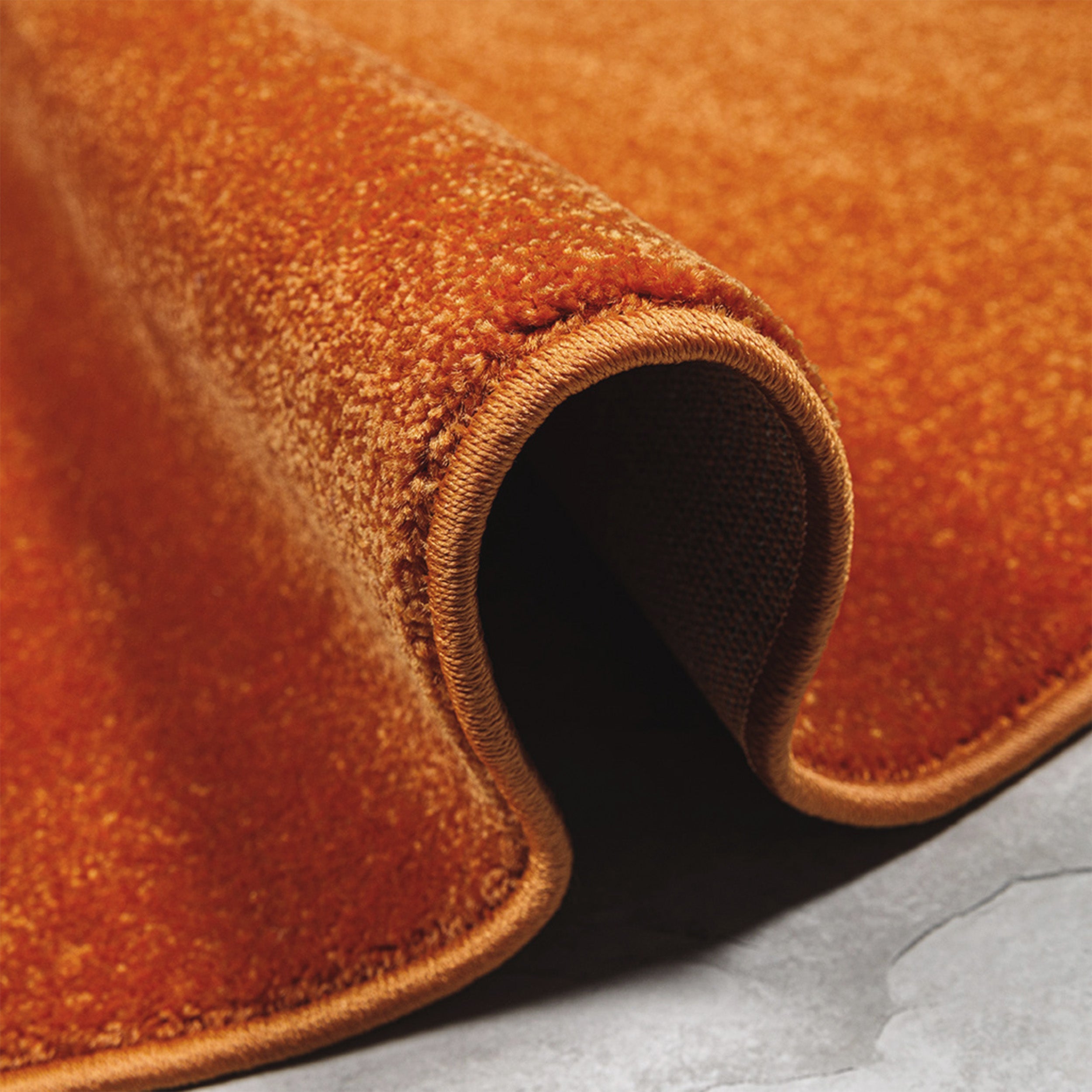 Gros plan d'un tapis orange terre cuite, partiellement enroulé, montrant sa texture dense et son bord ourlé sur un sol gris