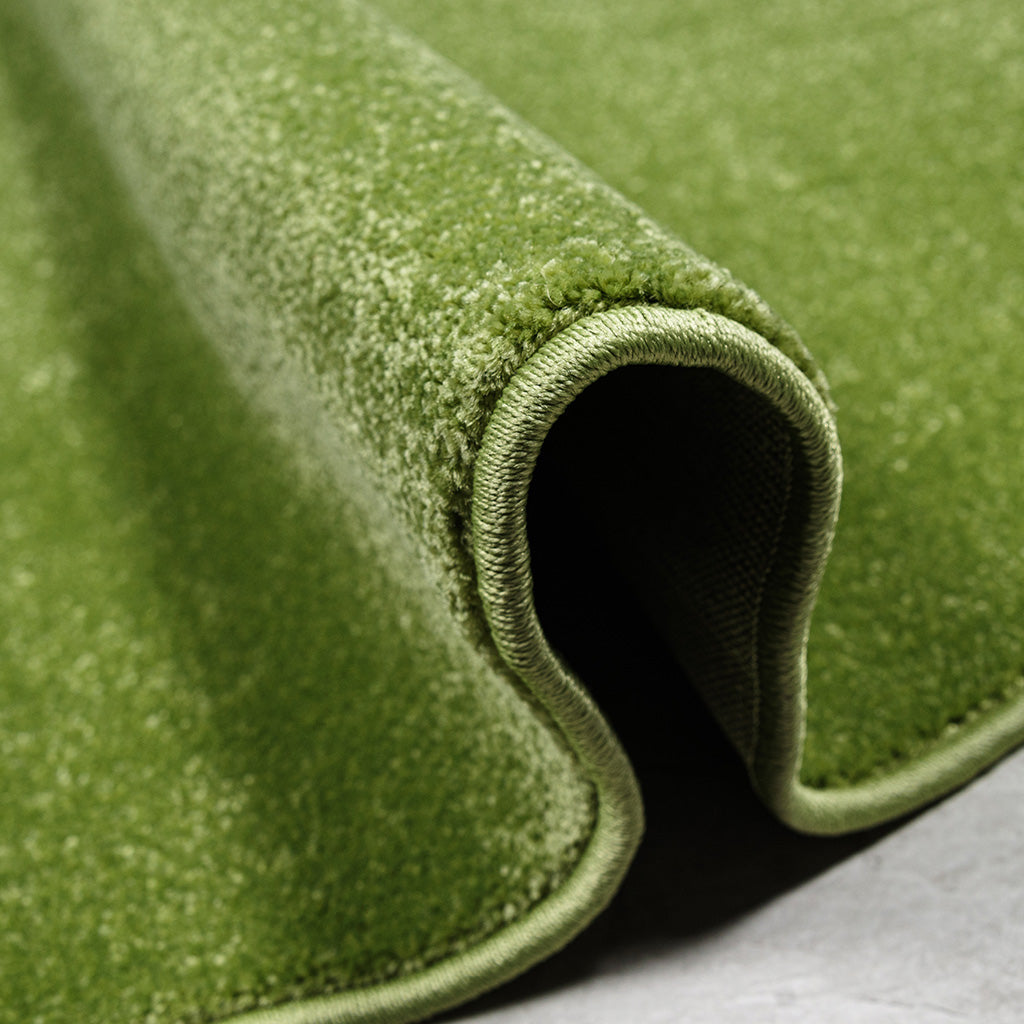 Détail d'un tapis vert mousse, à la texture moelleuse et aux bords surpiqués, roulé sur une surface grise