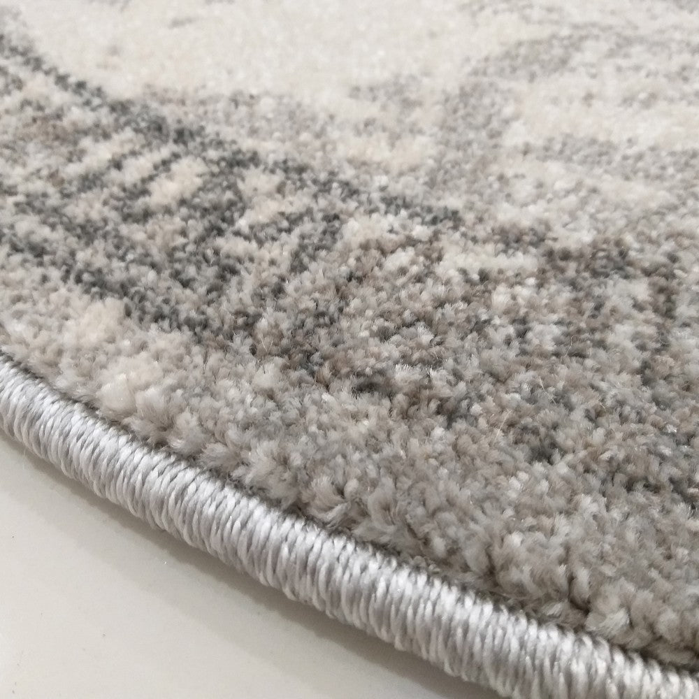 Gros plan sur un tapis moelleux aux teintes gris clair, blanc cassé et gris foncé, avec un motif abstrait texturé. Son bord est finement cousu