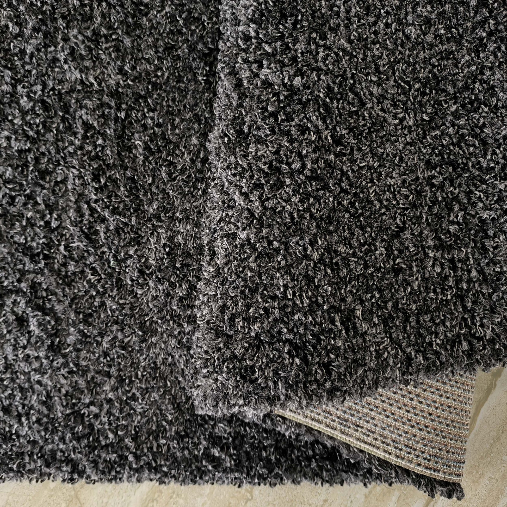 Tapis shaggy gris anthracite chiné aux poils longs et bouclés, son envers tissé et un sol beige sont visibles