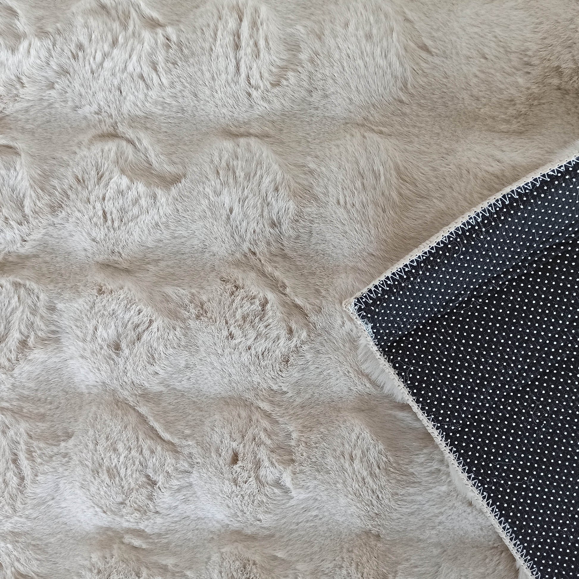 Tapis moelleux en fausse fourrure beige clair, avec un coin relevé montrant le dessous antidérapant noir à pois blancs