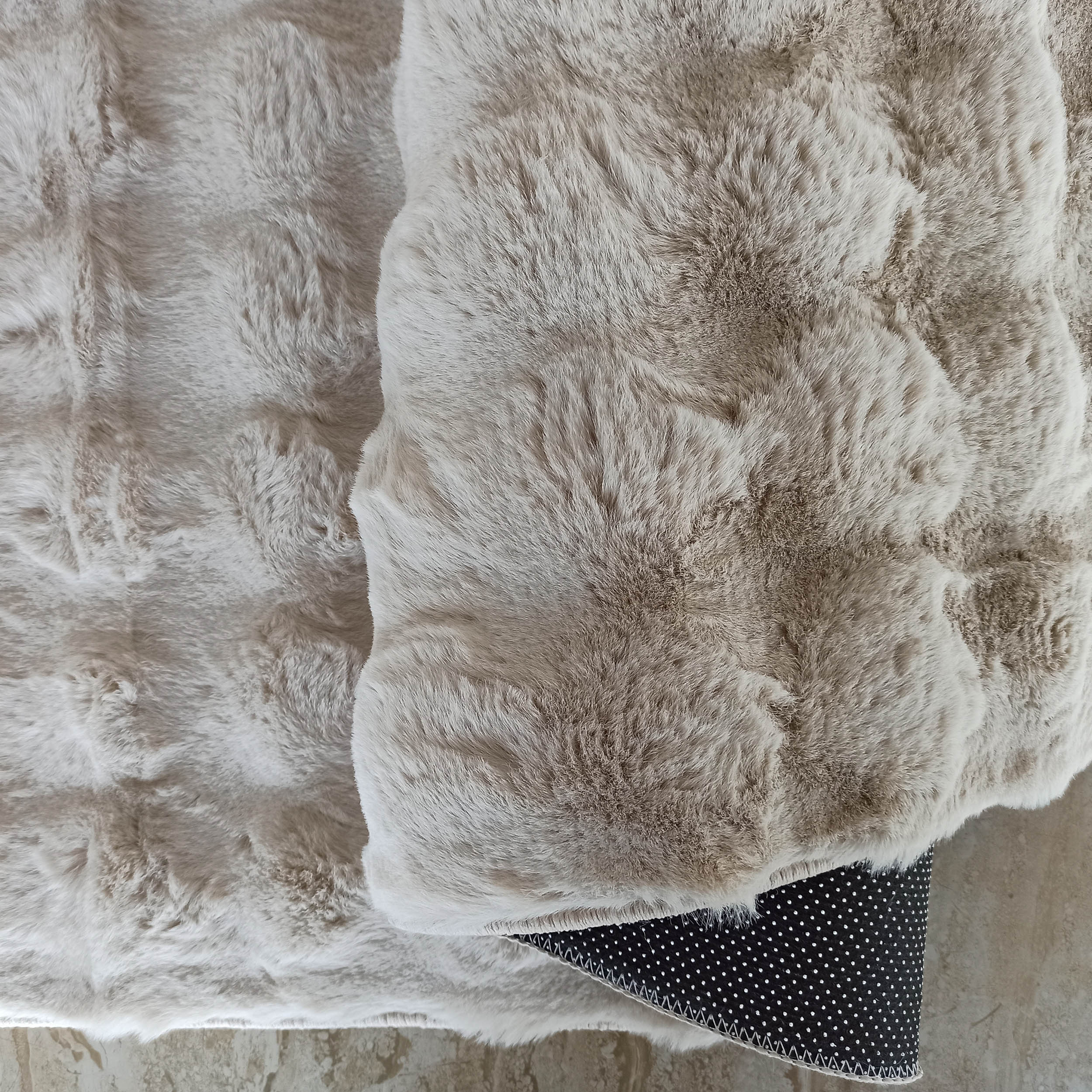 Tapis moelleux à poils longs beige clair, partiellement plié laissant apparaître son envers noir à petits pois blancs sur un sol carrelé clair