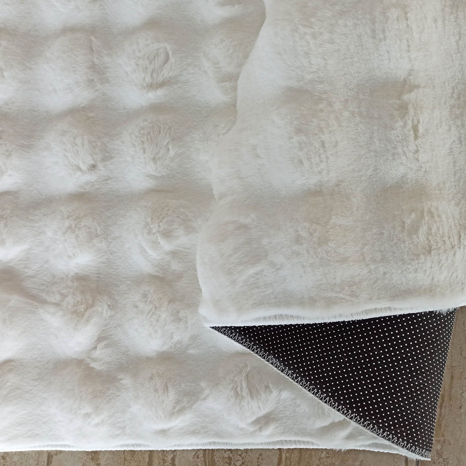 Tapis blanc cassé à poils longs, texture moelleuse. Un coin relevé montre le dessous noir à pois antidérapants blancs