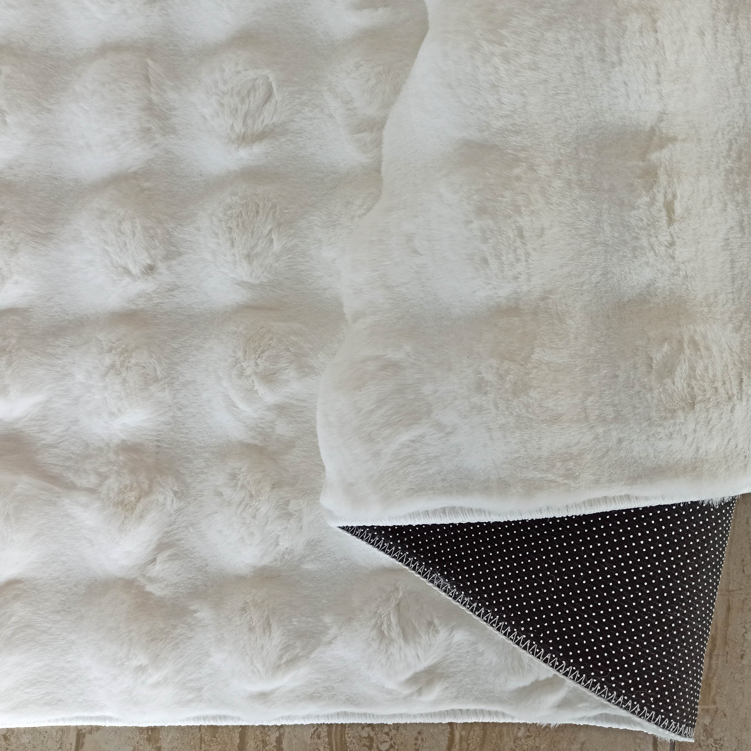 Tapis blanc cassé à poils longs, texture moelleuse. Un coin relevé montre le dessous noir à pois antidérapants blancs