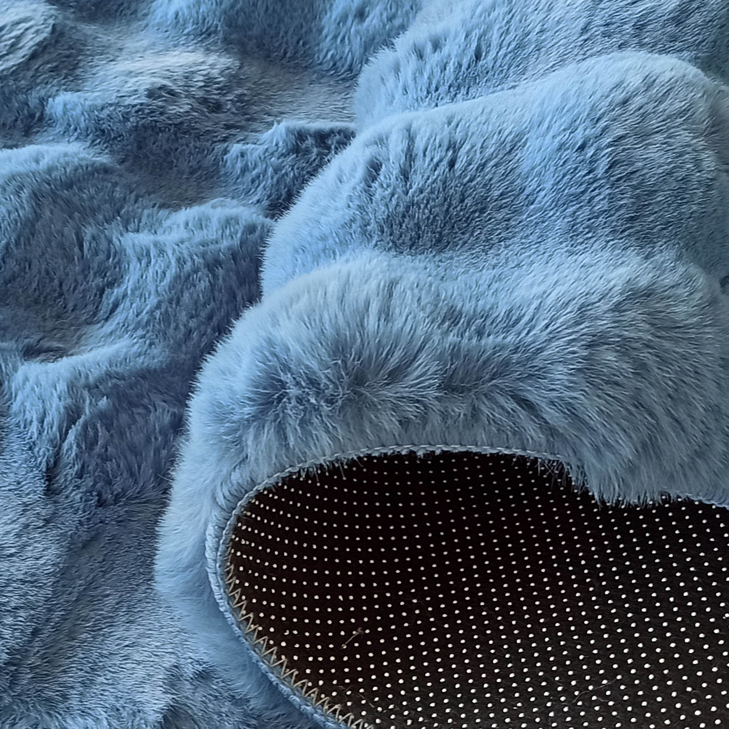 Tapis bleu ciel à poils longs et texture douce, avec un dessous noir à pois blancs antidérapants visible sur le côté