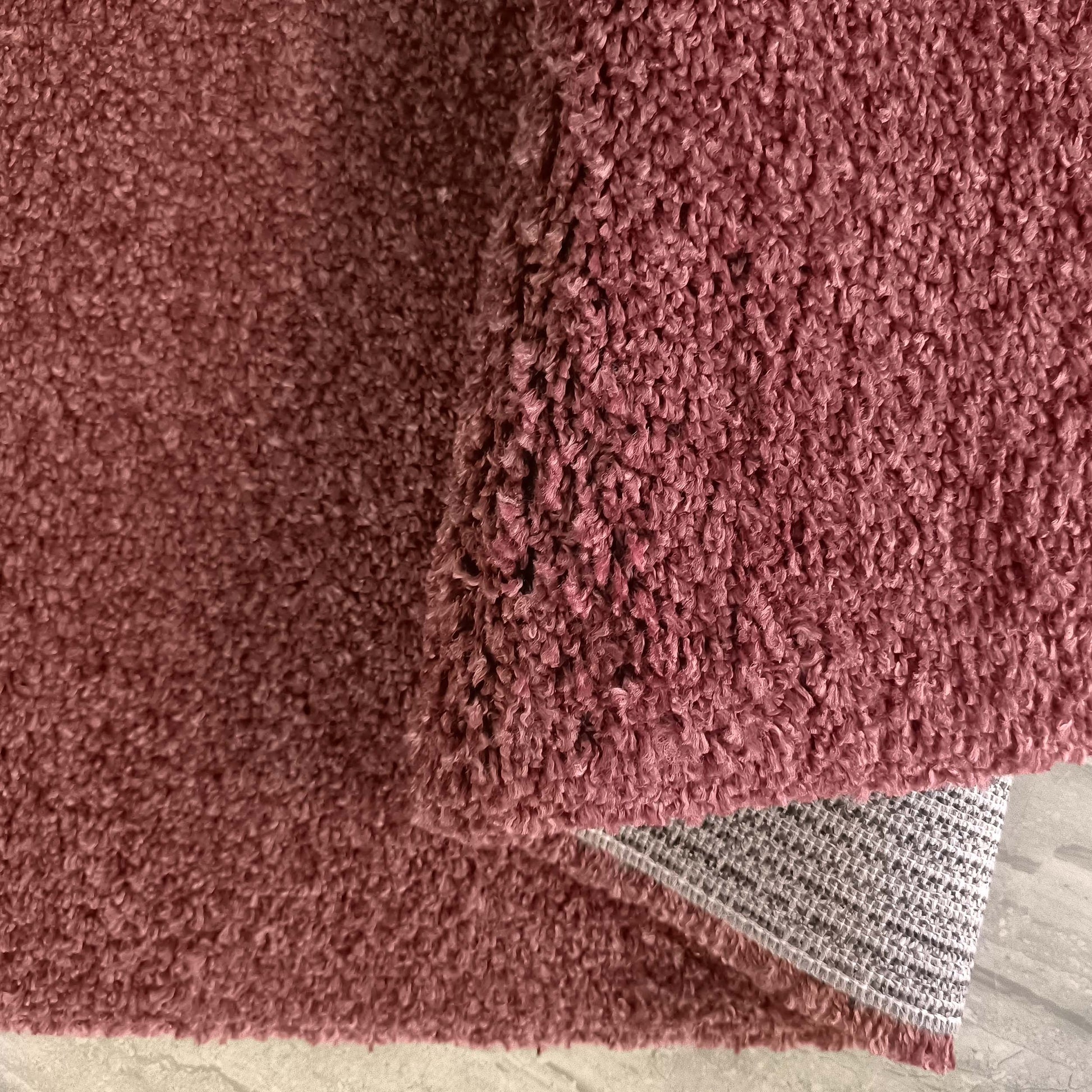 Tapis rose poudré à poils longs et texture douce, une partie est repliée laissant voir le dos antidérapant gris