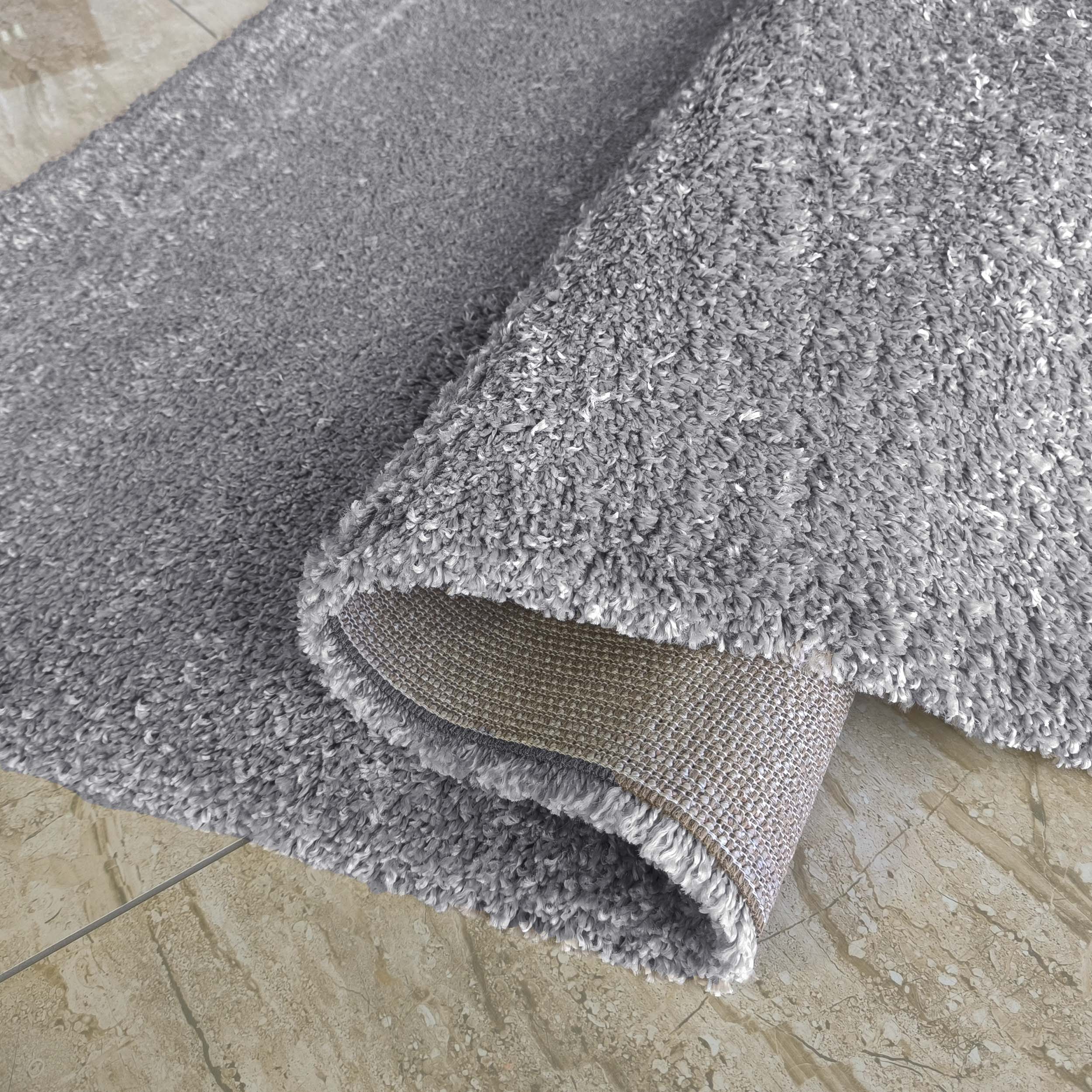 Tapis gris à poils longs, partiellement roulé sur un sol carrelé beige. Le dos du tapis révèle un tissage marron clair