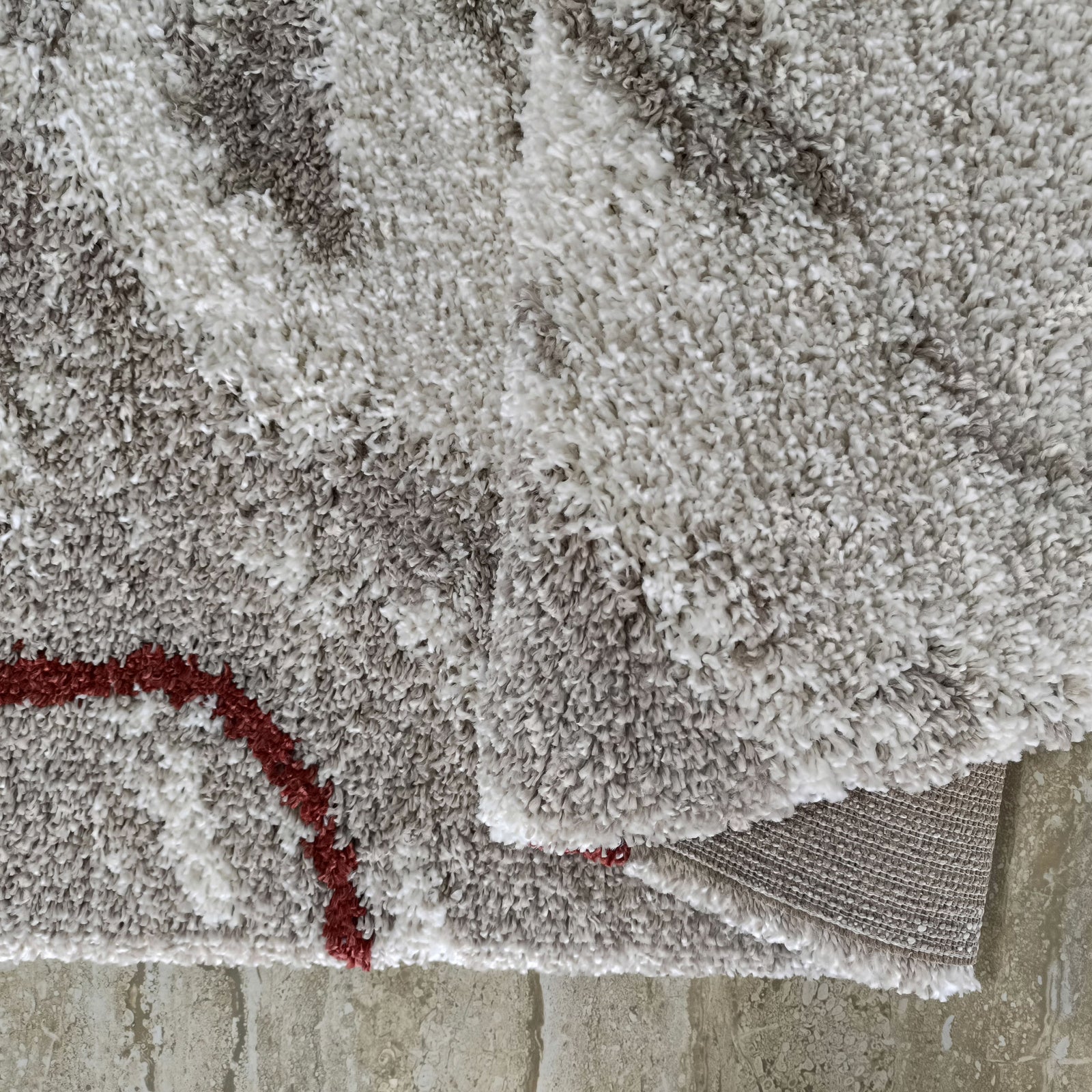 Tapis moelleux gris et blanc aux motifs abstraits, avec une ligne rouge. Le tapis est partiellement plié montrant son envers tissé