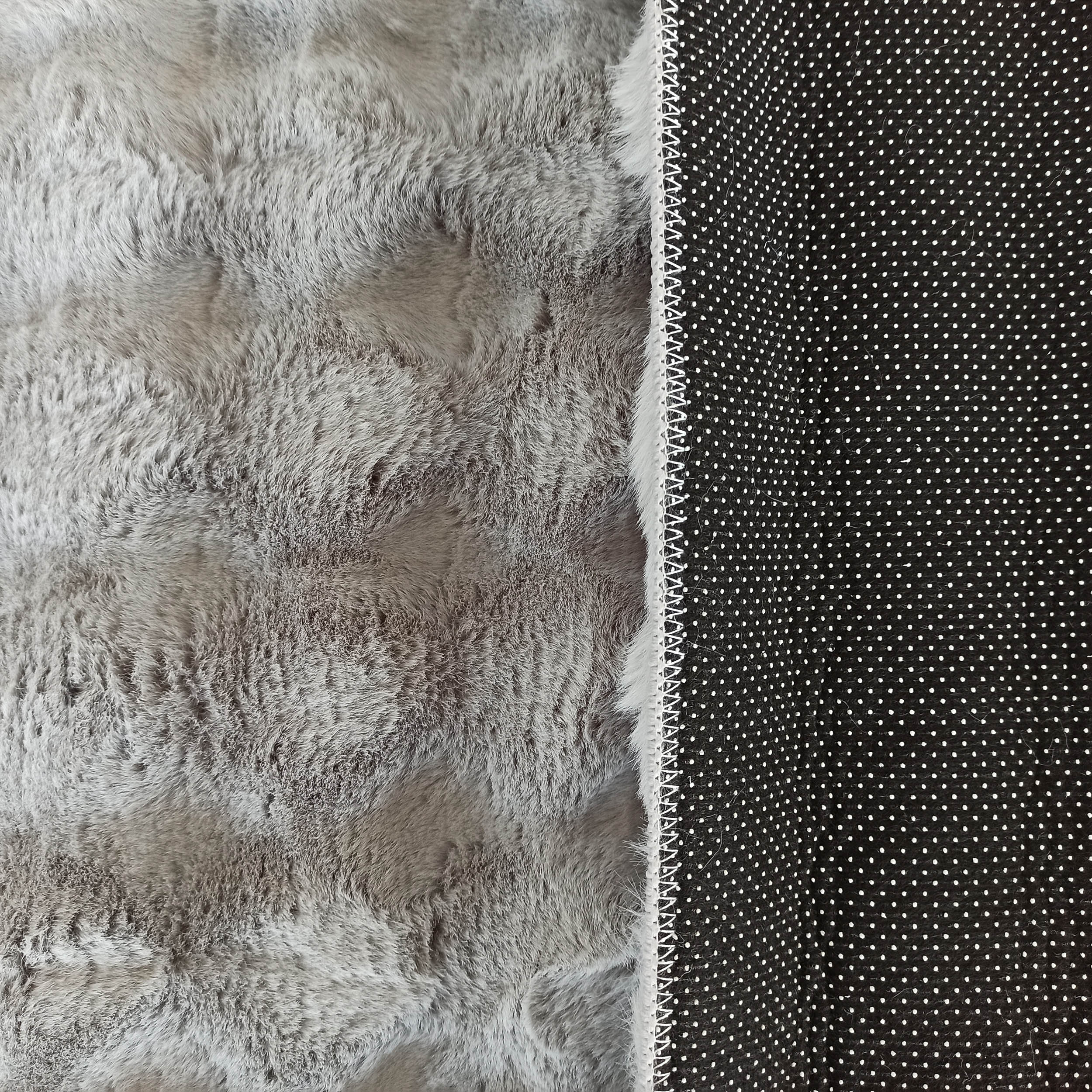 Tapis gris clair à poils longs, avec son envers noir parsemé de petits points blancs antidérapants et une couture blanche