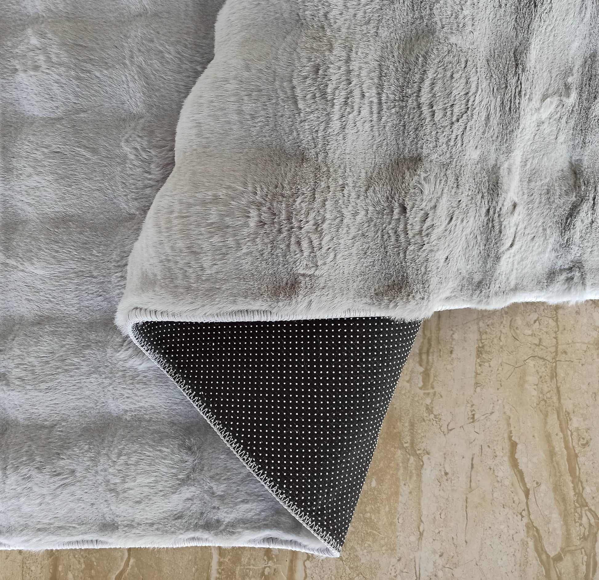 Tapis gris clair à la texture douce comme de la fourrure, dont un coin montre l'envers noir antidérapant à pois sur un sol en marbre beige