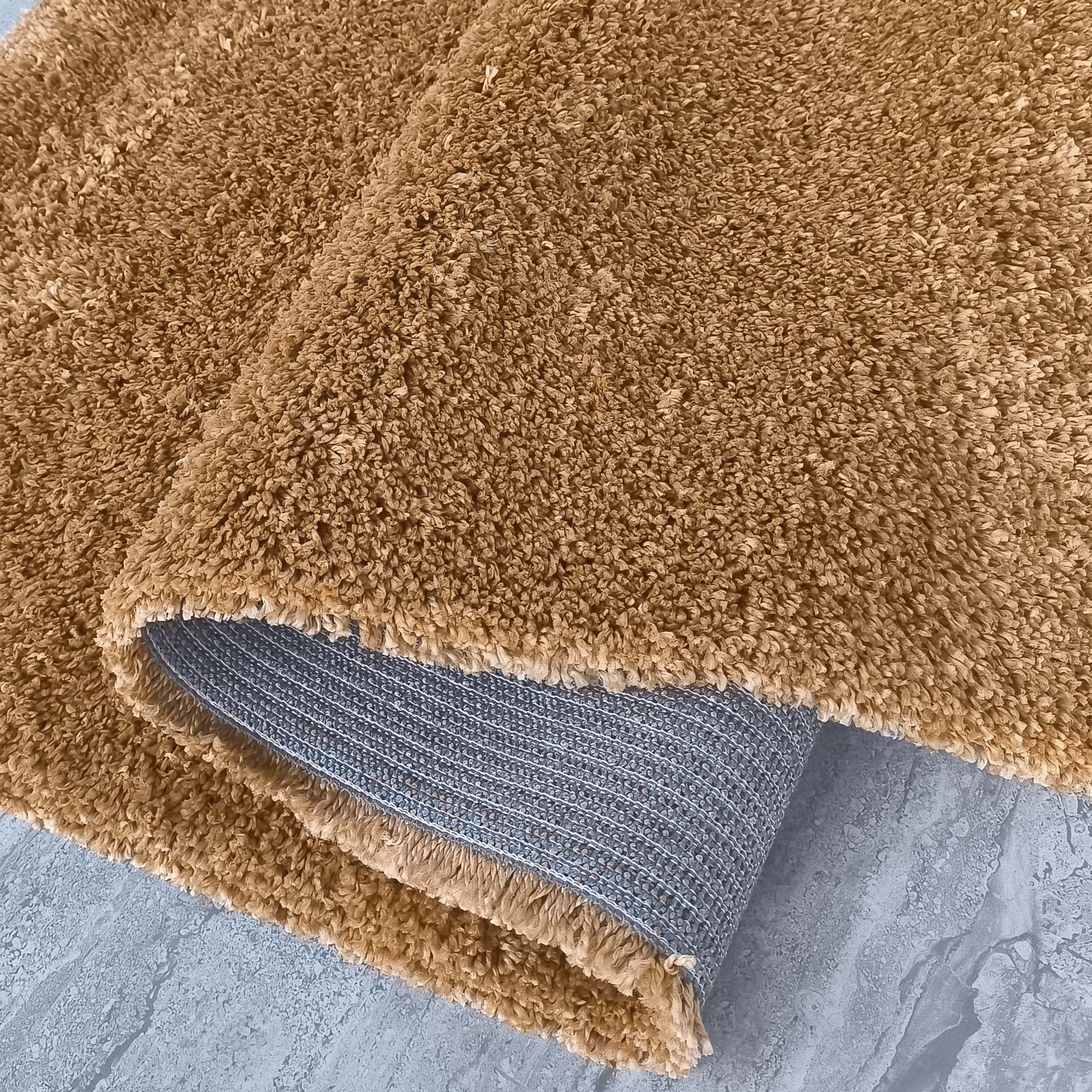 Tapis marron clair à poils longs, partiellement roulé pour montrer son envers gris quadrillé, sur un sol texturé gris