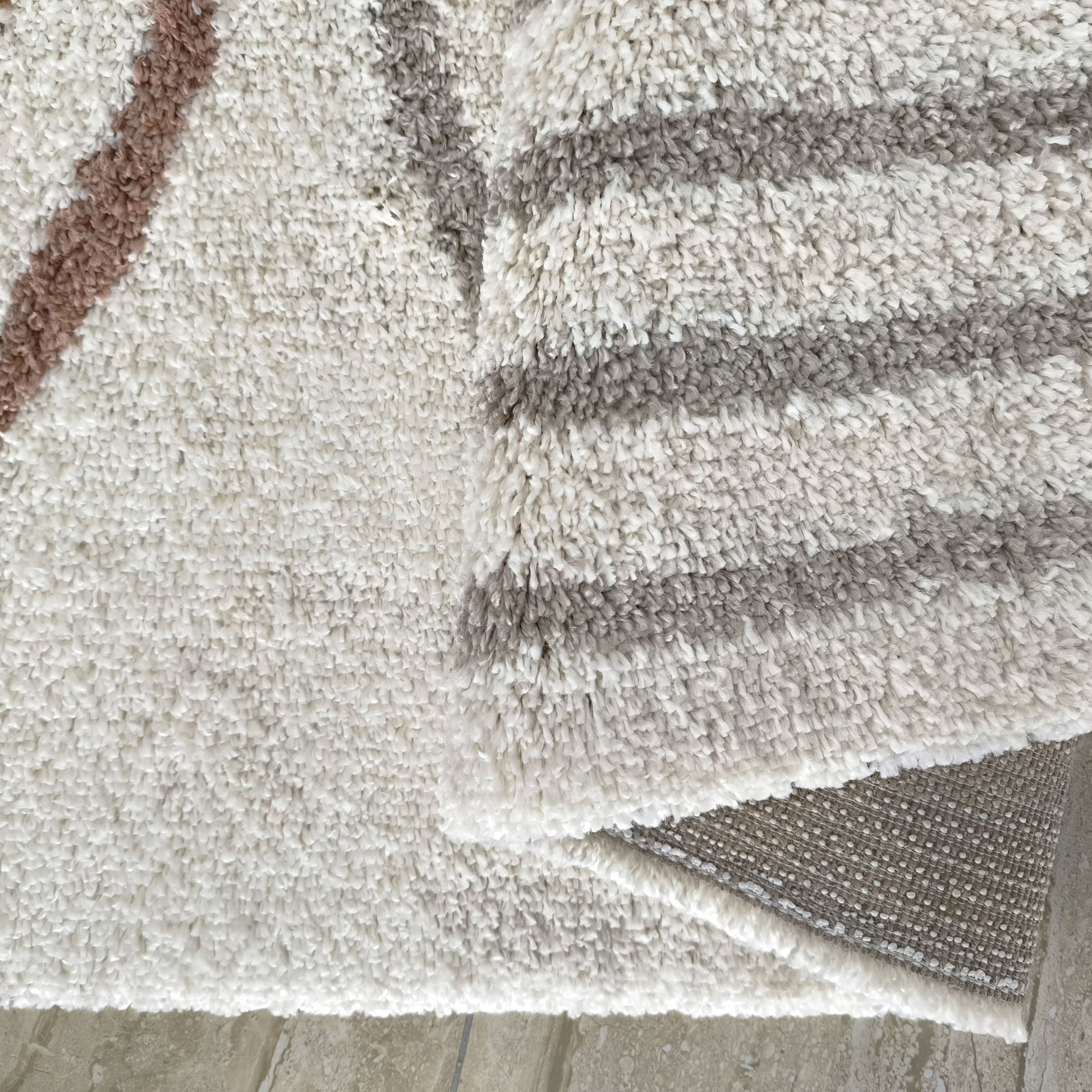 Tapis à poils longs blanc cassé, avec des motifs diagonaux brun rosé et des rayures grises. Le revers tissé du tapis est visible