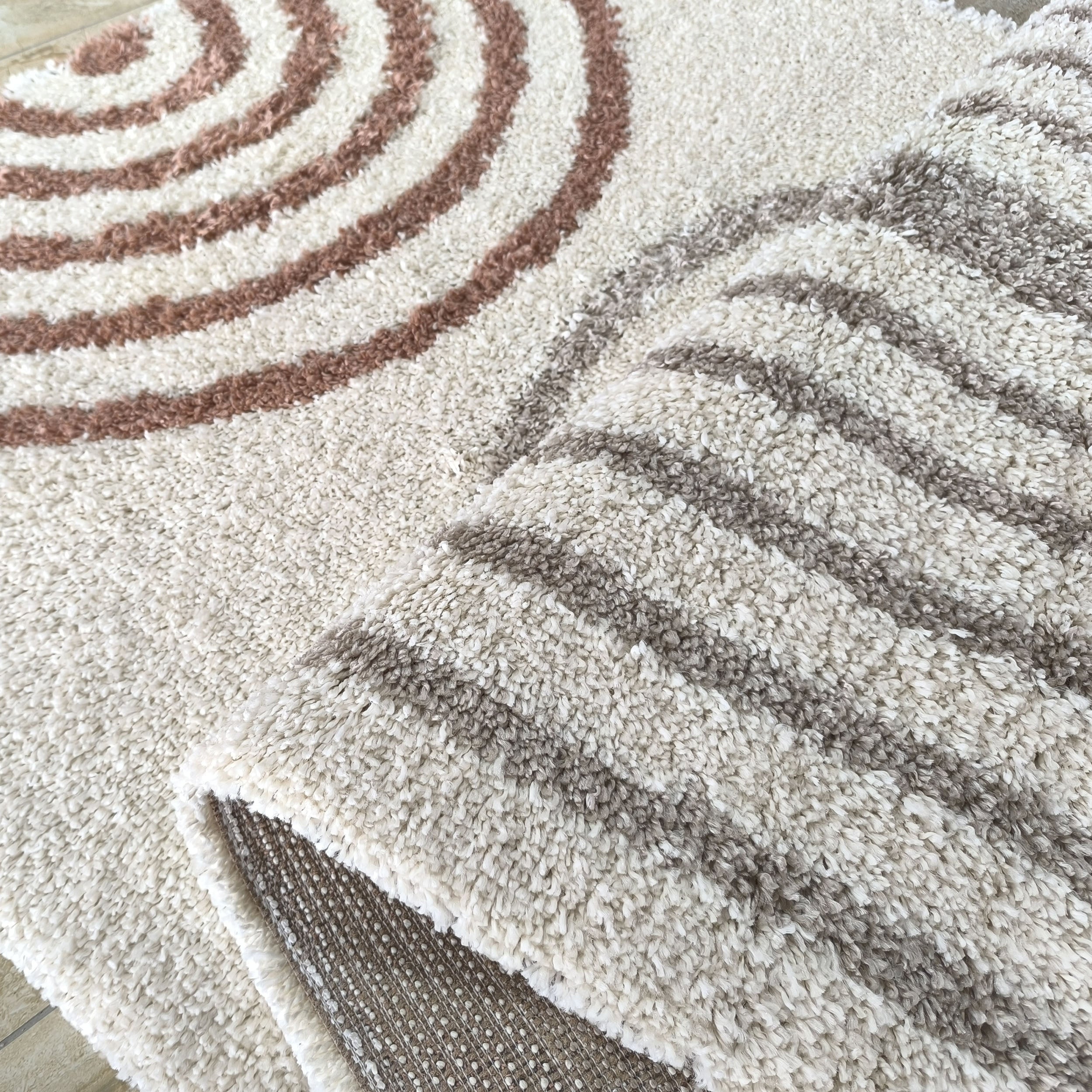 Tapis shaggy épais crème à poils longs avec motifs de cercles concentriques marron. Le tapis est replié, révélant son envers pointillé