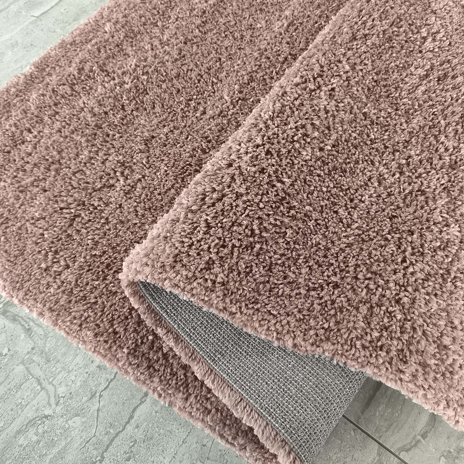 Tapis rose poudré à texture douce et bouclée, partiellement roulé montrant son envers tissé gris sur un sol clair