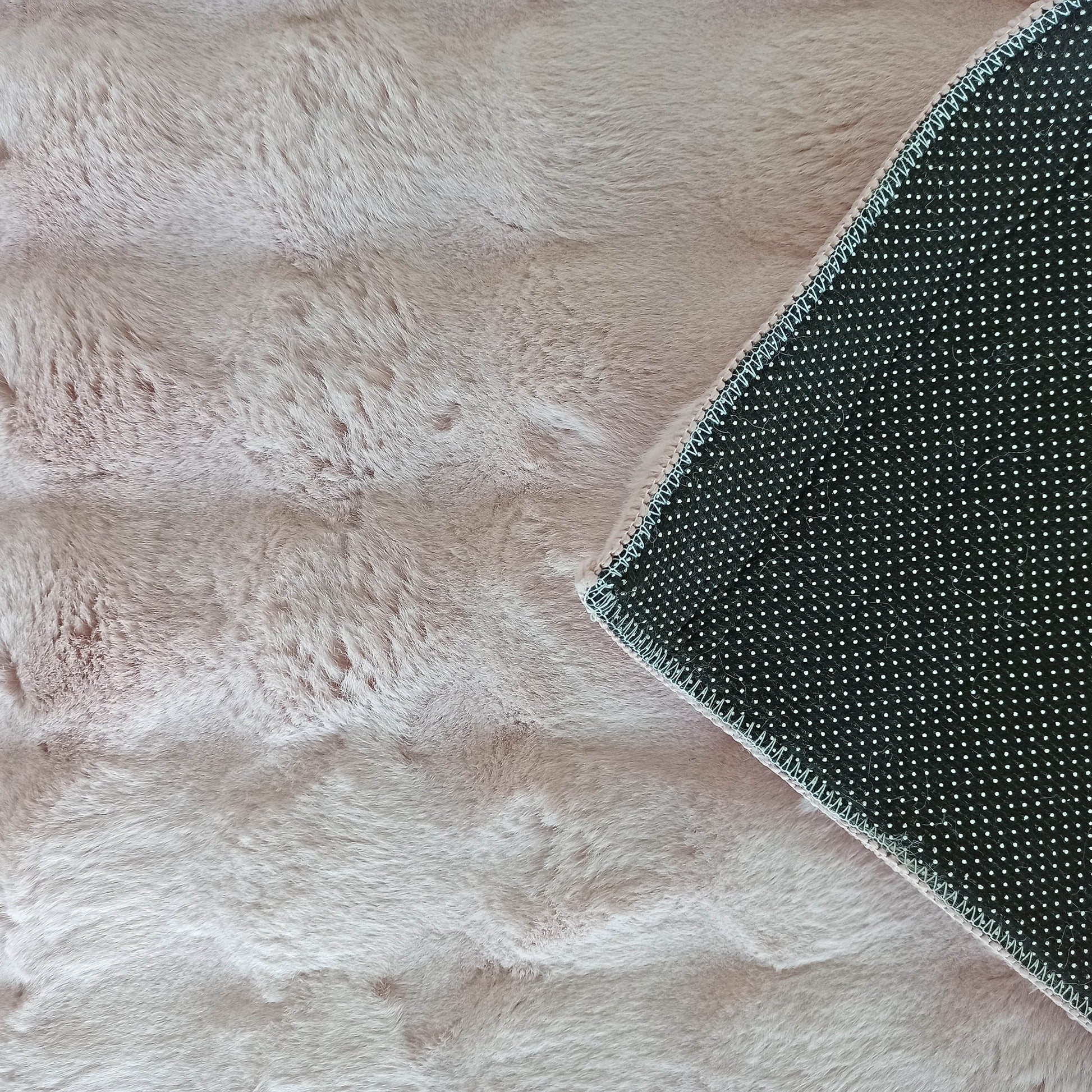 Tapis rose clair à poils longs, doux et moelleux, dont un coin est replié, montrant le dessous noir à pois blancs et des coutures claires