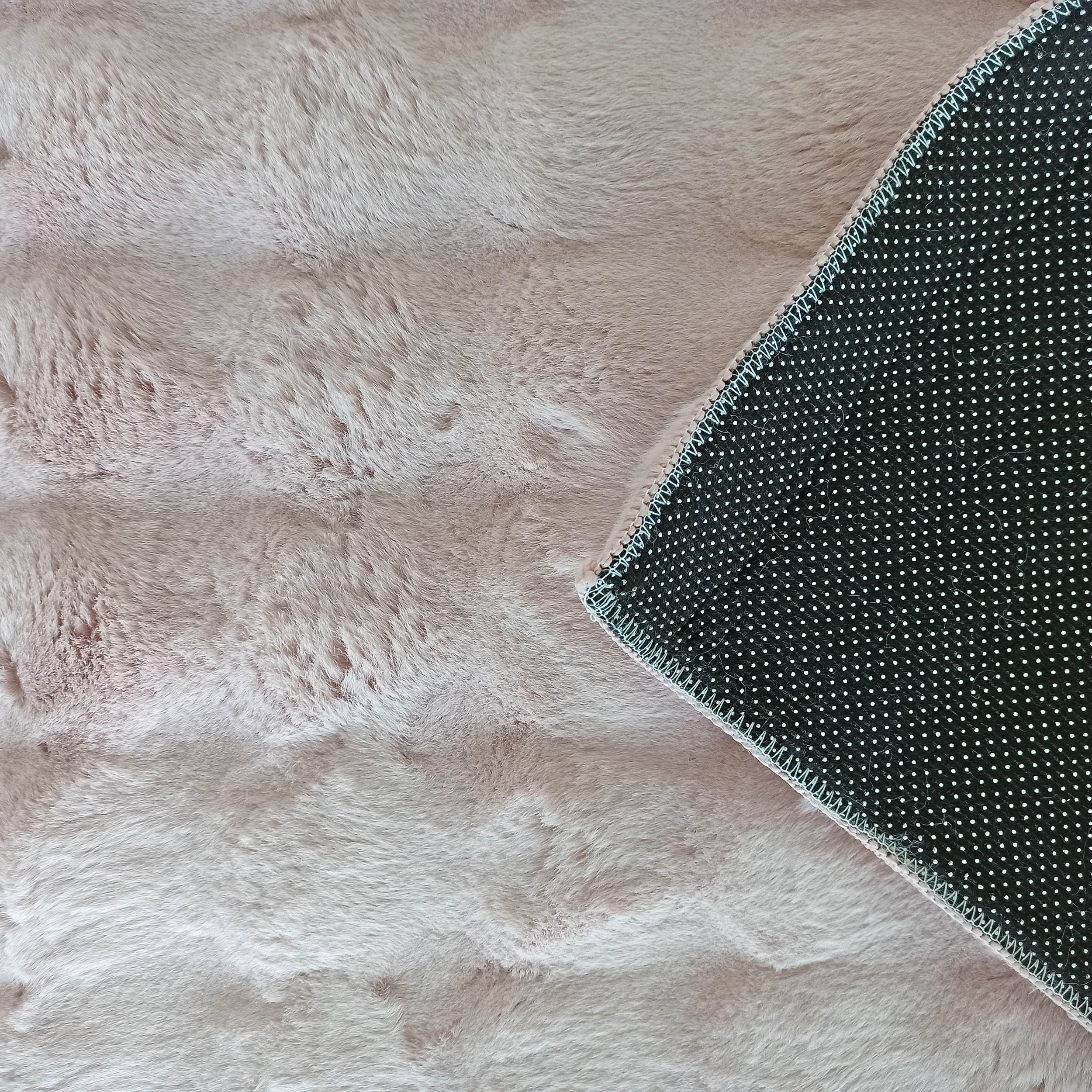Tapis rose clair à poils longs, doux et moelleux, dont un coin est replié, montrant le dessous noir à pois blancs et des coutures claires
