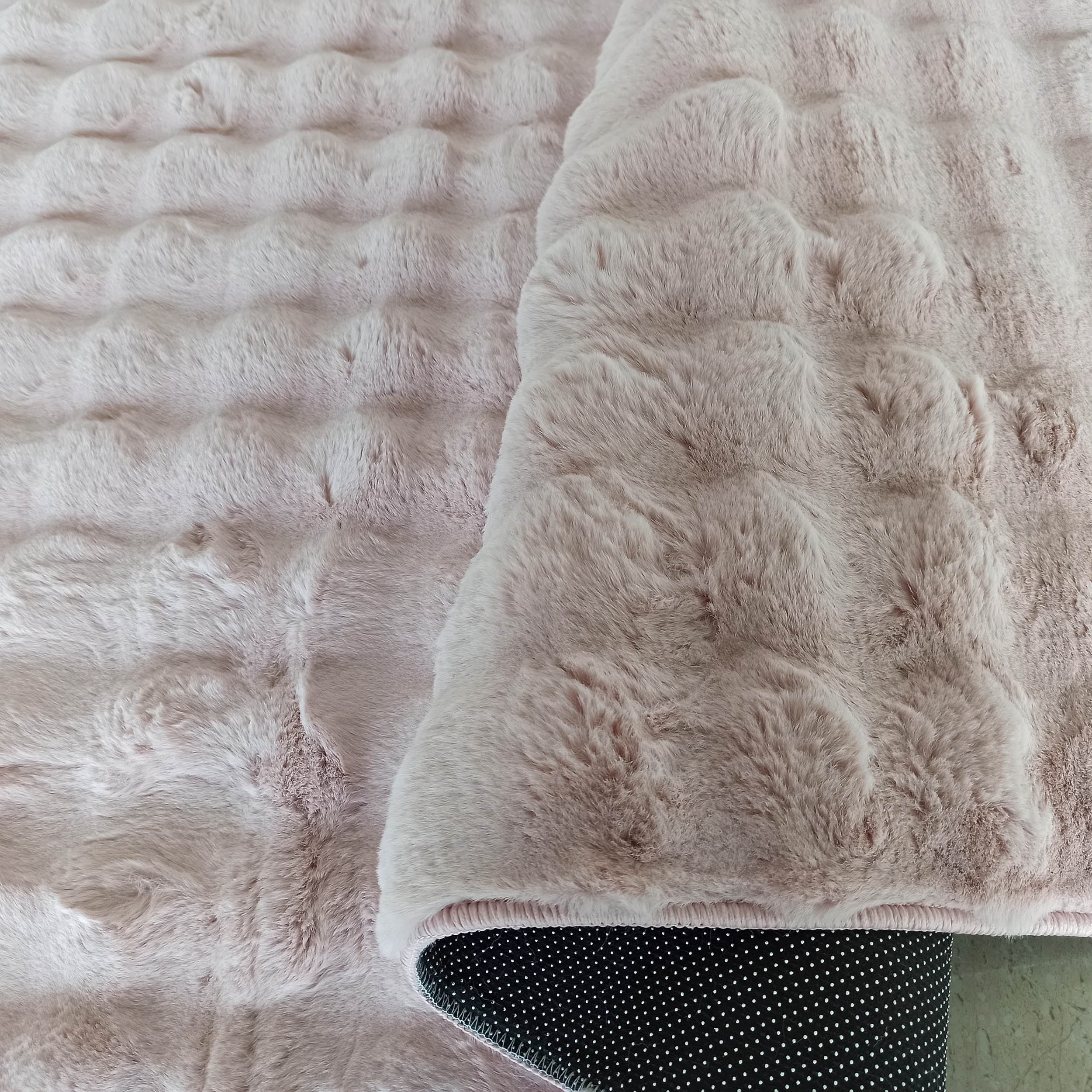 Tapis rose pâle et moelleux avec une surface en relief ondulée, le dessous noir à pois blancs est visible