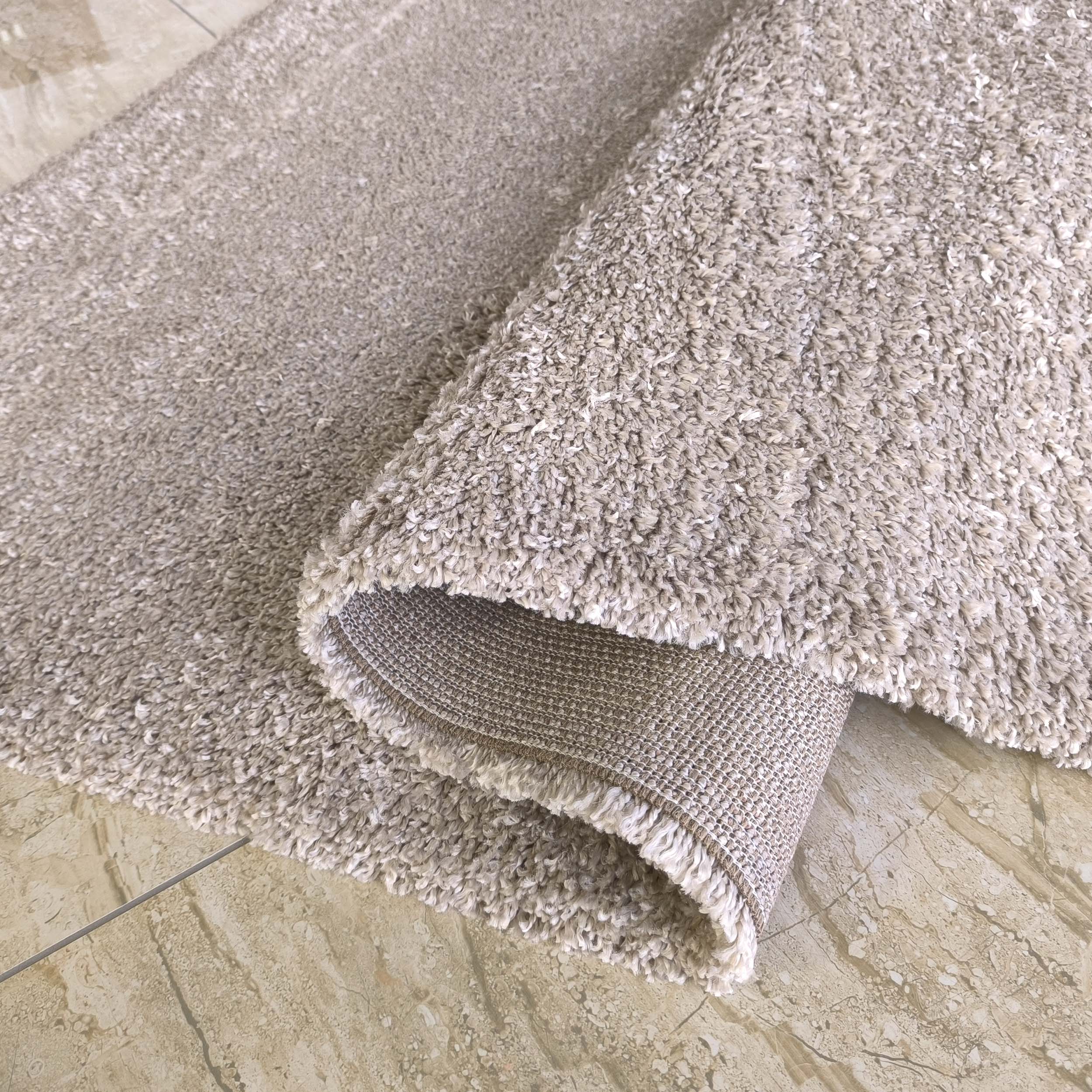 Tapis shaggy beige clair à mèches mélangées, enroulé pour montrer sa doublure tissée sur un sol carrelé marbré