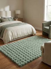Tapis rectangulaire vert sauge à motifs carrés en relief, moelleux et doux, posé sur un parquet en bois clair dans une chambre