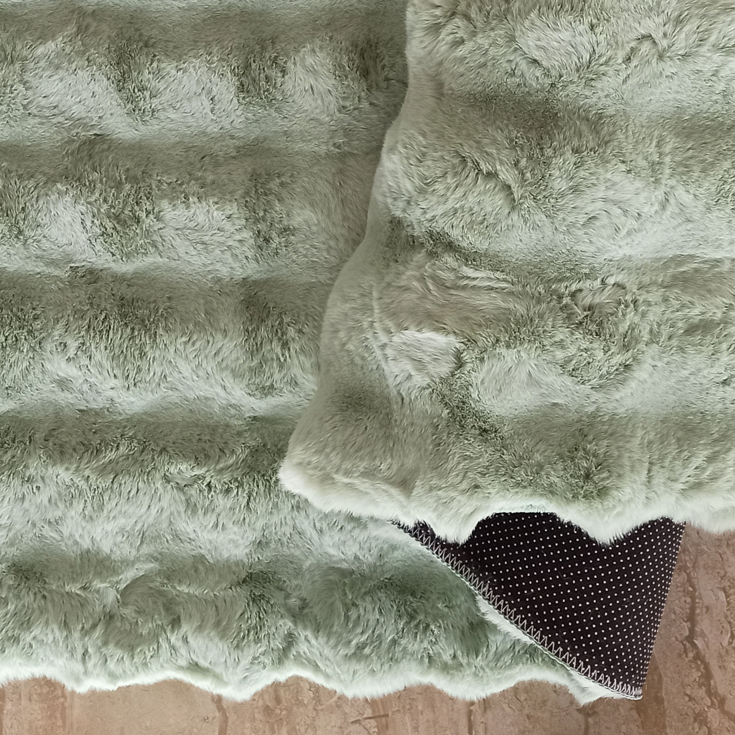 Tapis vert sauge à poils longs créant des motifs texturés, replié sur un sol clair pour montrer son envers noir à pois blancs