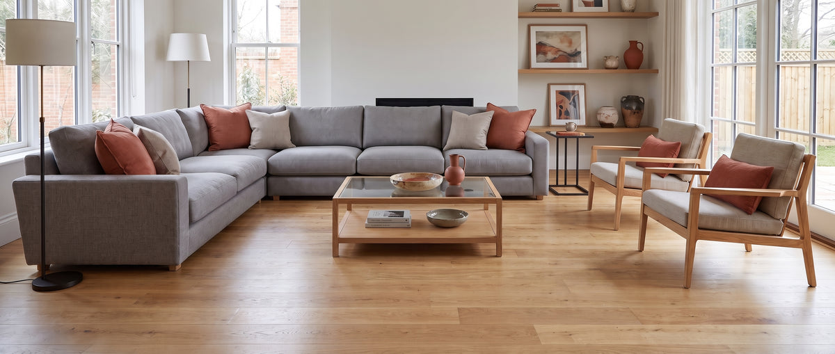 Dans un salon lumineux au sol en bois nu, un grand canapé gris avec coussins terracotta et des fauteuils, sans tapis