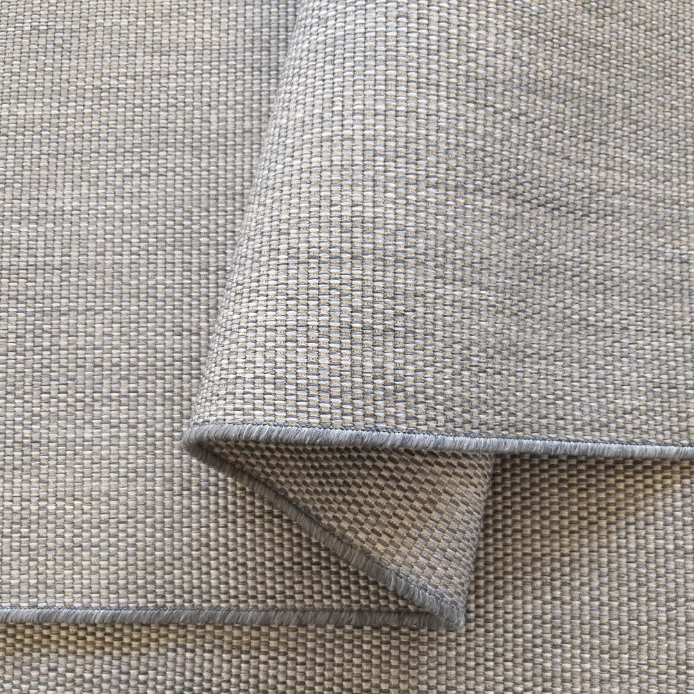 Tapis gris clair à la texture tissée serrée formant de petits carrés, avec un pan plié révélant un bord cousu gris foncé