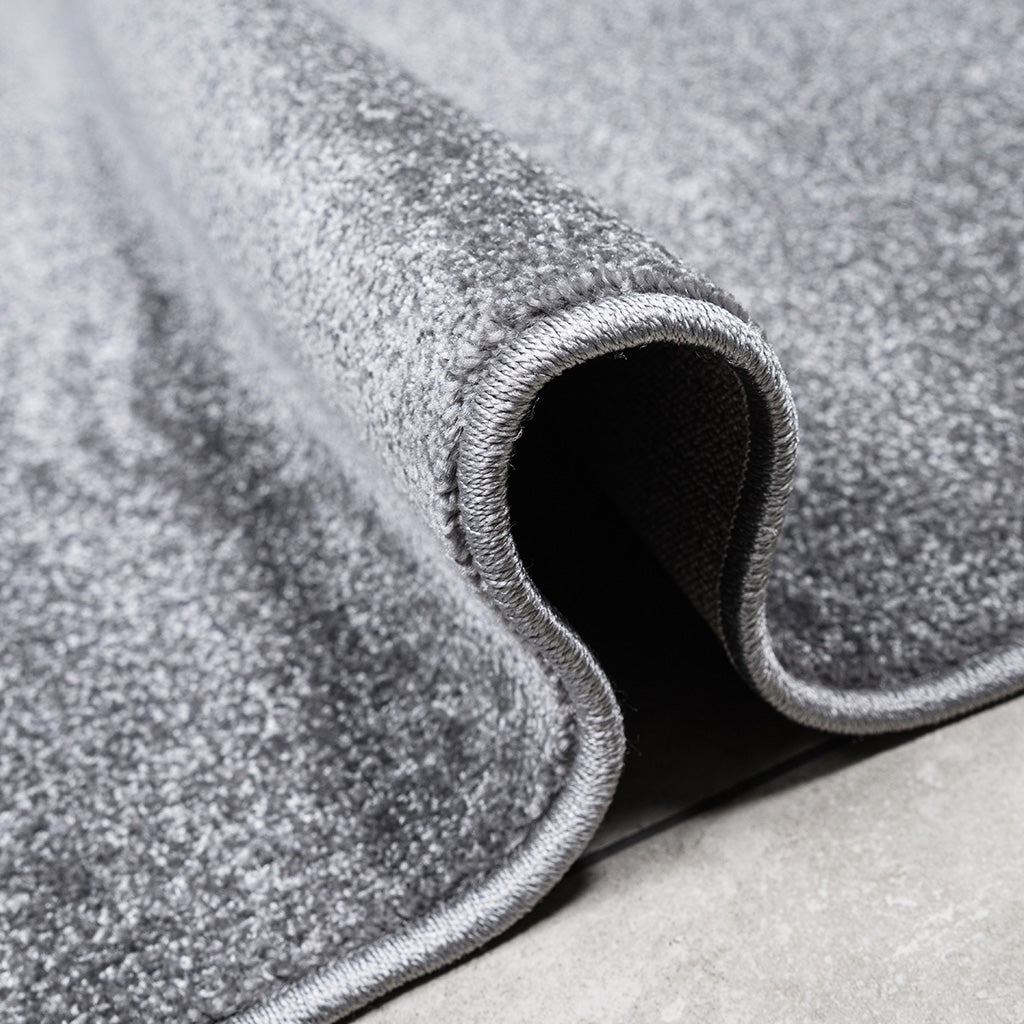 Gros plan d'un tapis gris chiné, à la texture douce, partiellement roulé, montrant sa bordure cousue