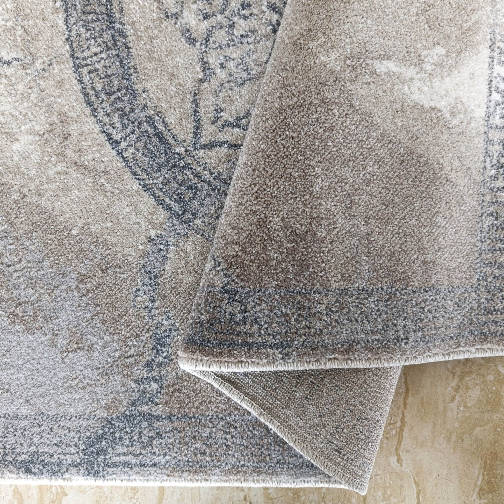 Tapis texturé aux motifs complexes beiges et gris foncé, partiellement roulé sur un sol carrelé beige marbré