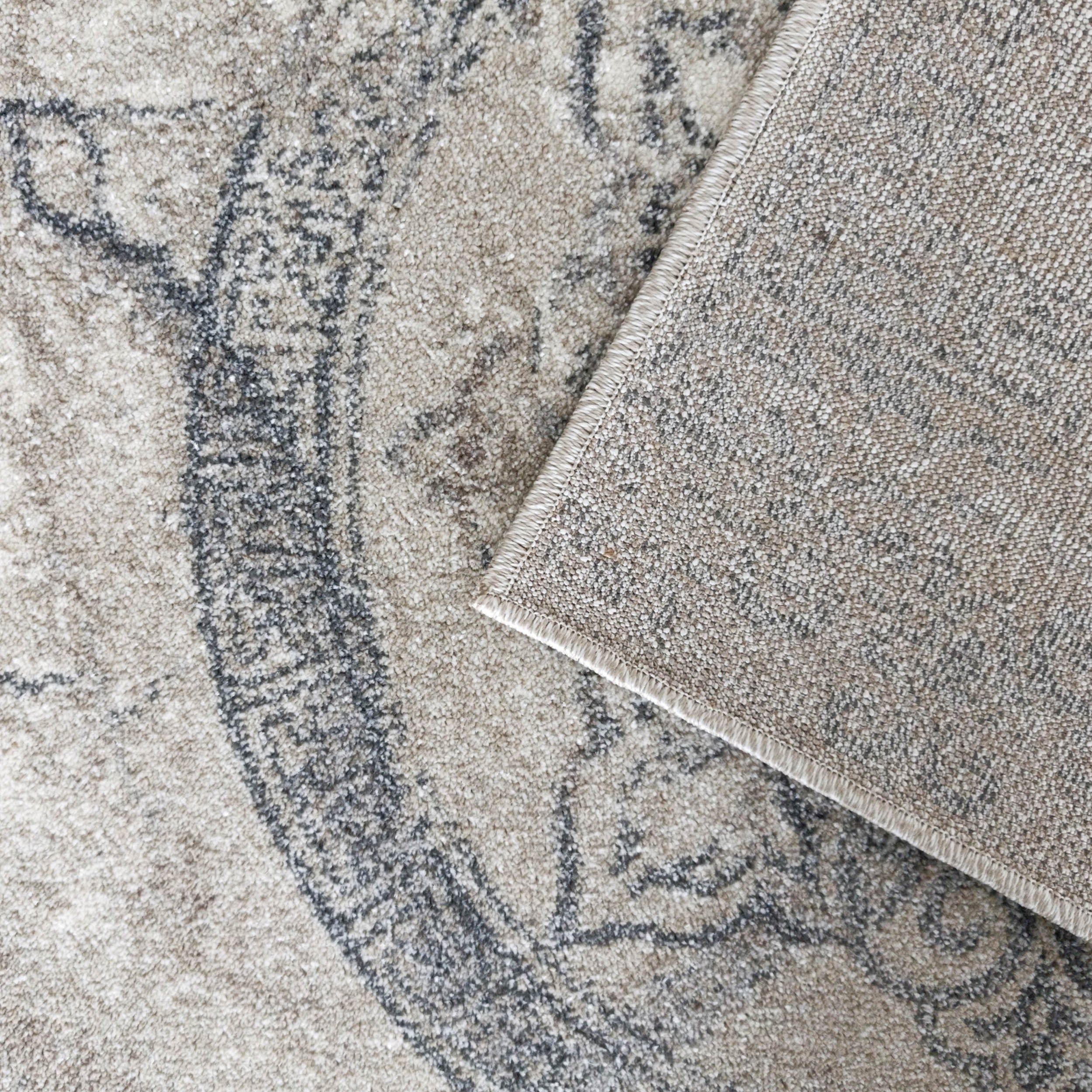 Tapis doux beige avec des motifs ornementaux gris foncé, un coin est relevé montrant l'envers texturé en tissu chiné beige