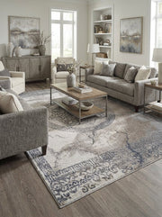 Tapis rectangulaire tissé à motifs traditionnels délavés, mêlant des teintes de beige, gris et bleu foncé, posé dans un salon