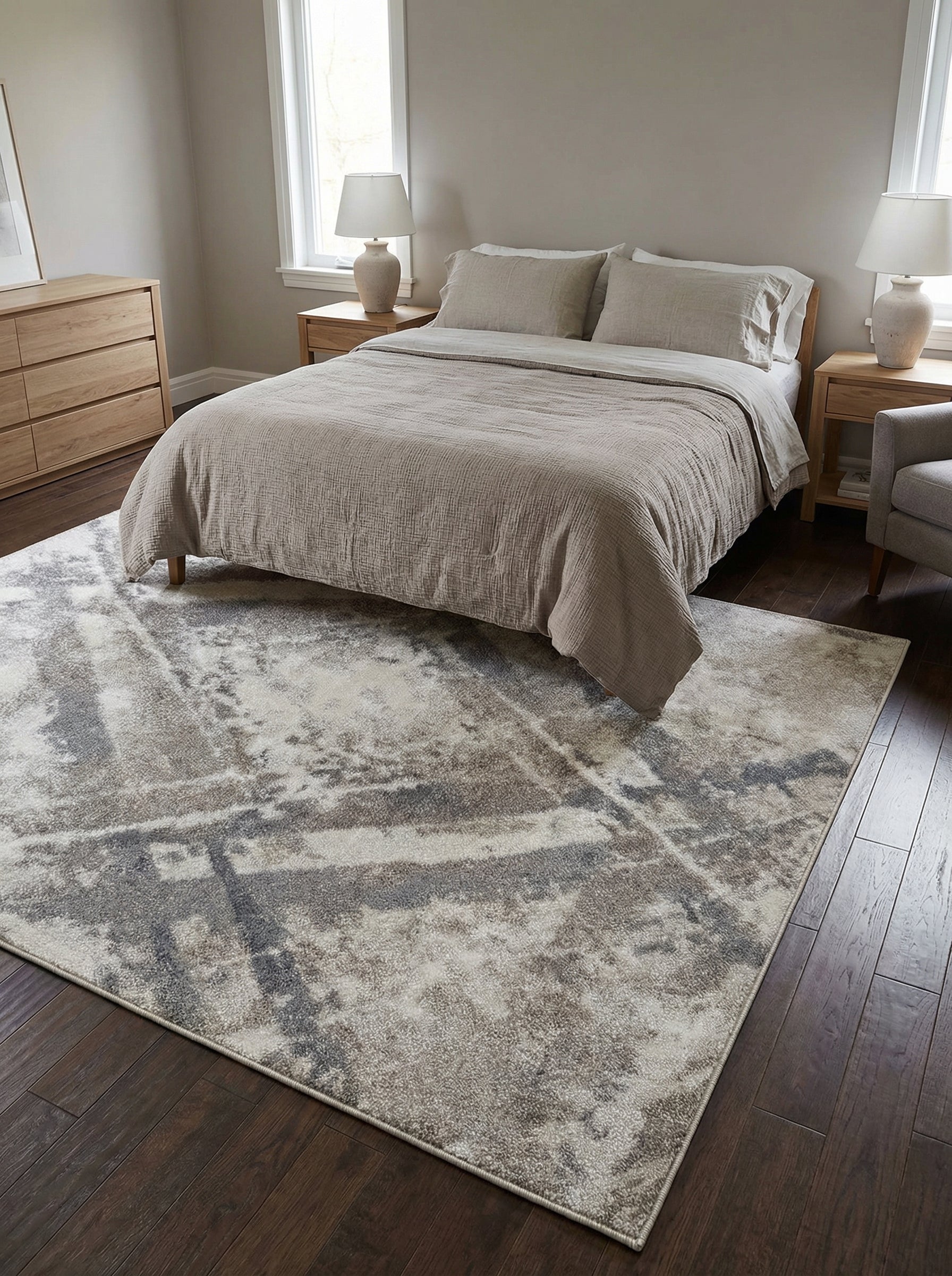 Tapis rectangulaire aux motifs abstraits gris clair, beige et gris foncé, évoquant des coups de pinceau diffus. Posé sur un plancher sombre dans une chambre moderne