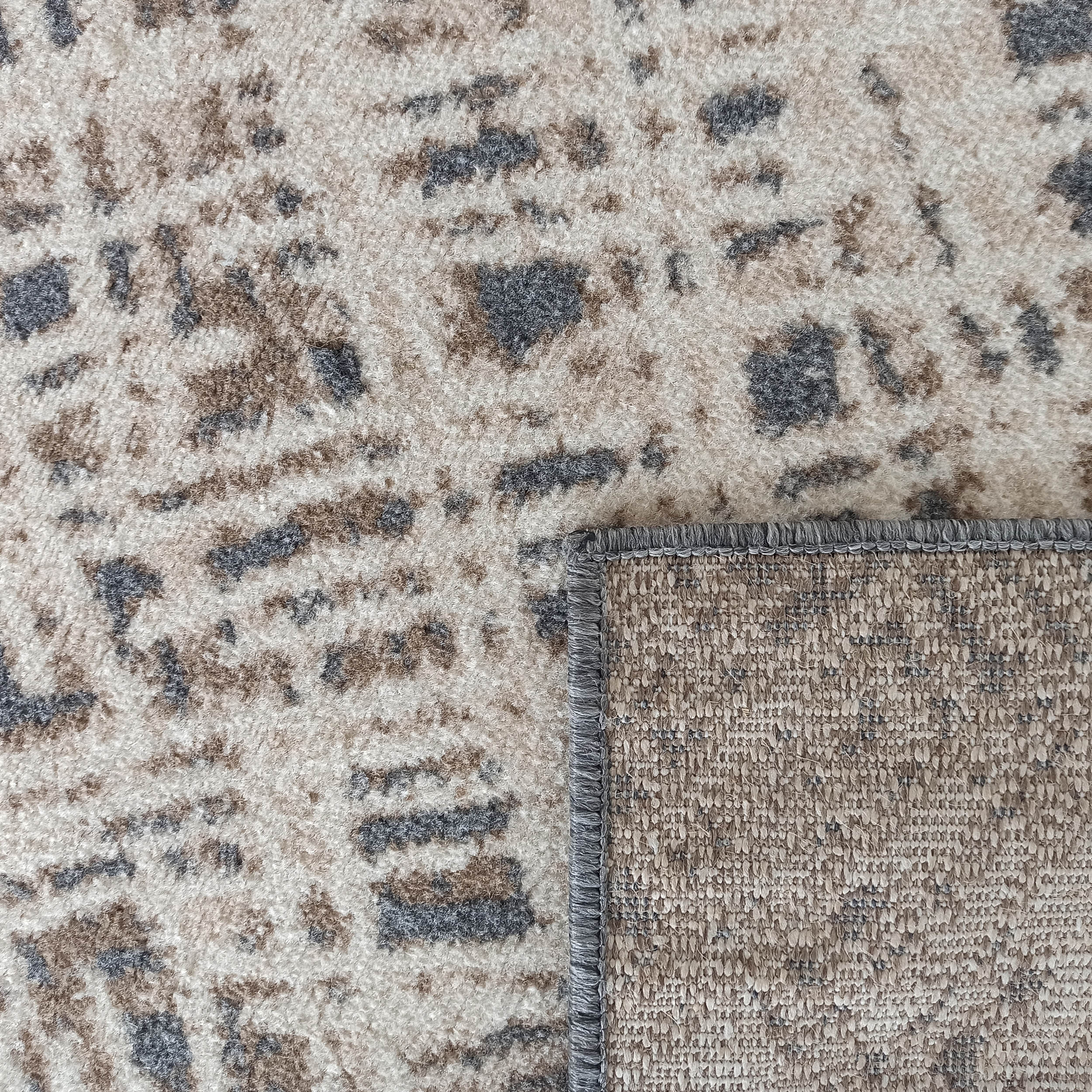 Tapis crème texturé avec motifs irréguliers marron et gris, montrant sa sous-face tissée beige et sa bordure grise