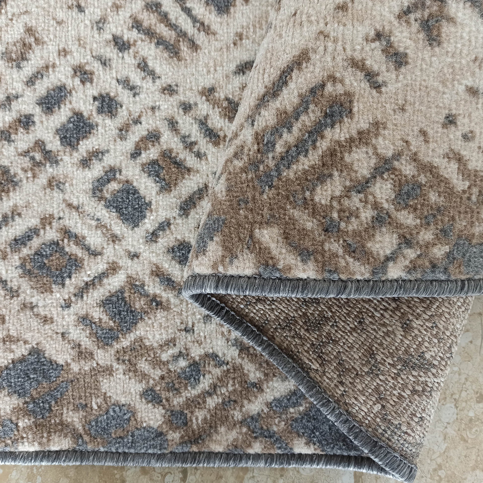 Tapis doux aux motifs géométriques irréguliers crème, gris et marron, partiellement replié, montrant son envers texturé