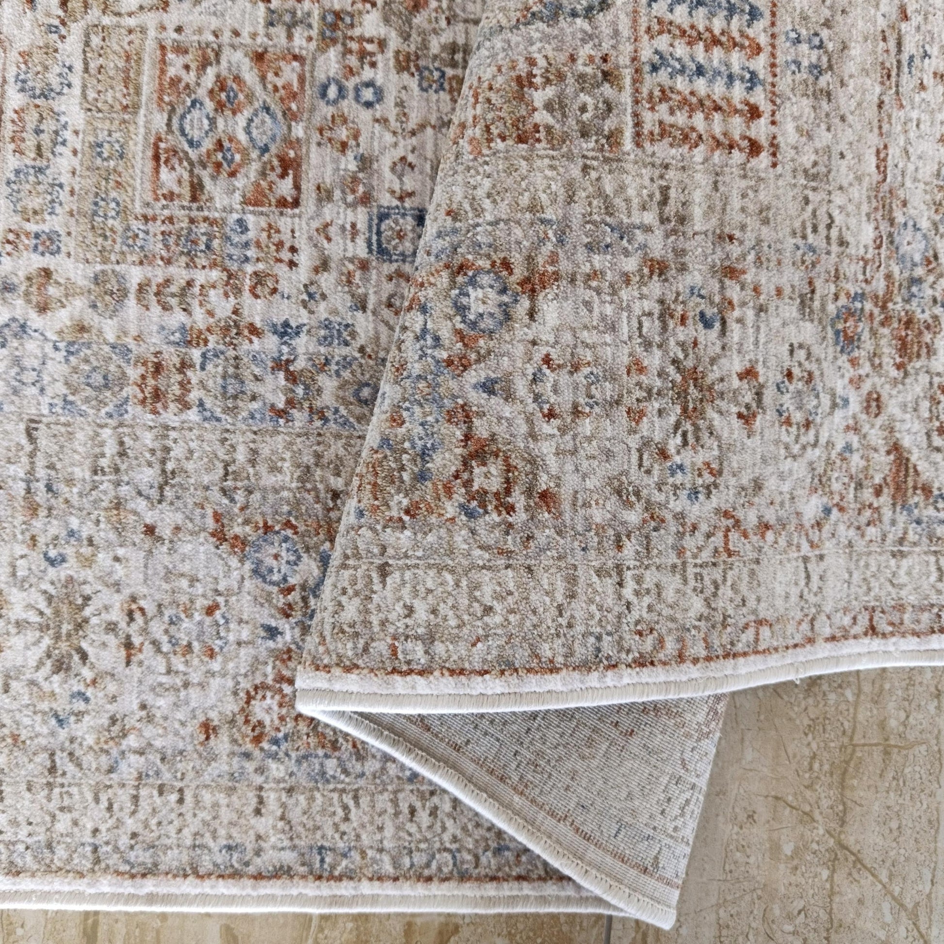 Tapis doux beige à motifs géométriques et floraux bleus et rouille, partiellement enroulé sur un sol clair marbré
