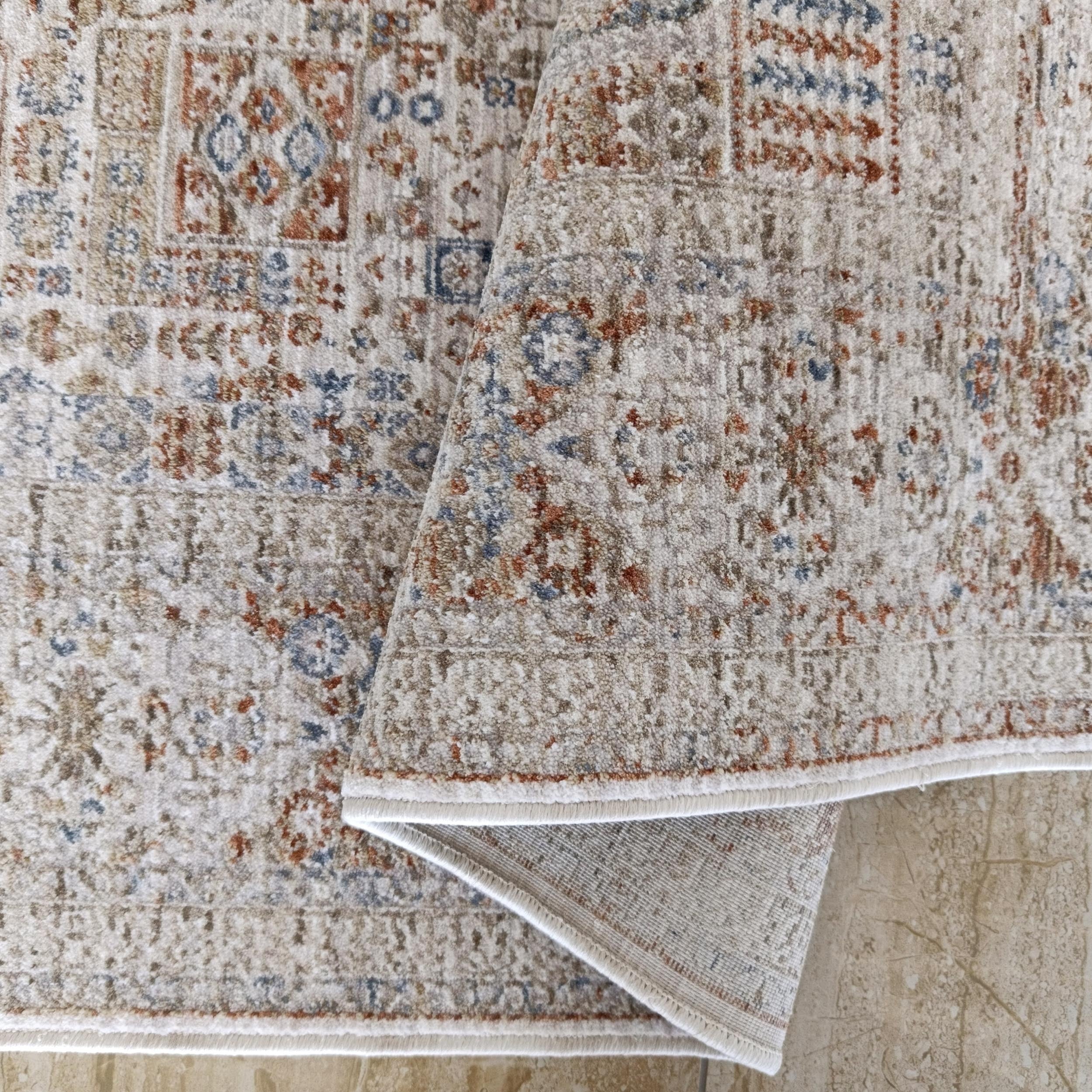 Tapis doux beige à motifs géométriques et floraux bleus et rouille, partiellement enroulé sur un sol clair marbré
