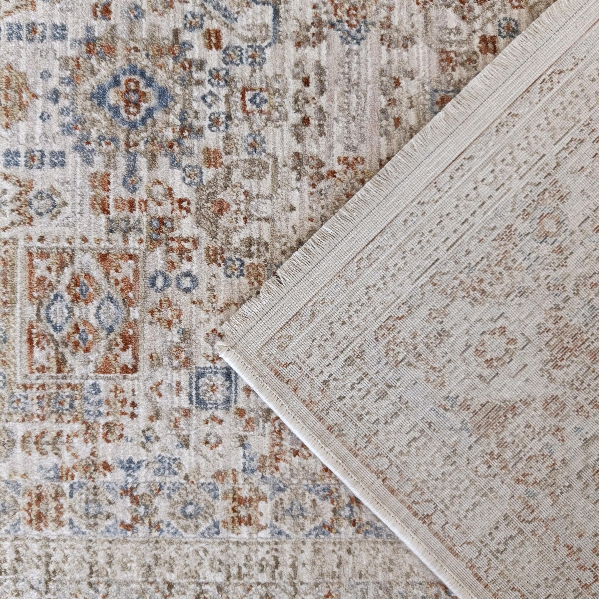 Tapis beige à motifs complexes rouille et bleu, un coin relevé révèle le revers tissé crème et les franges